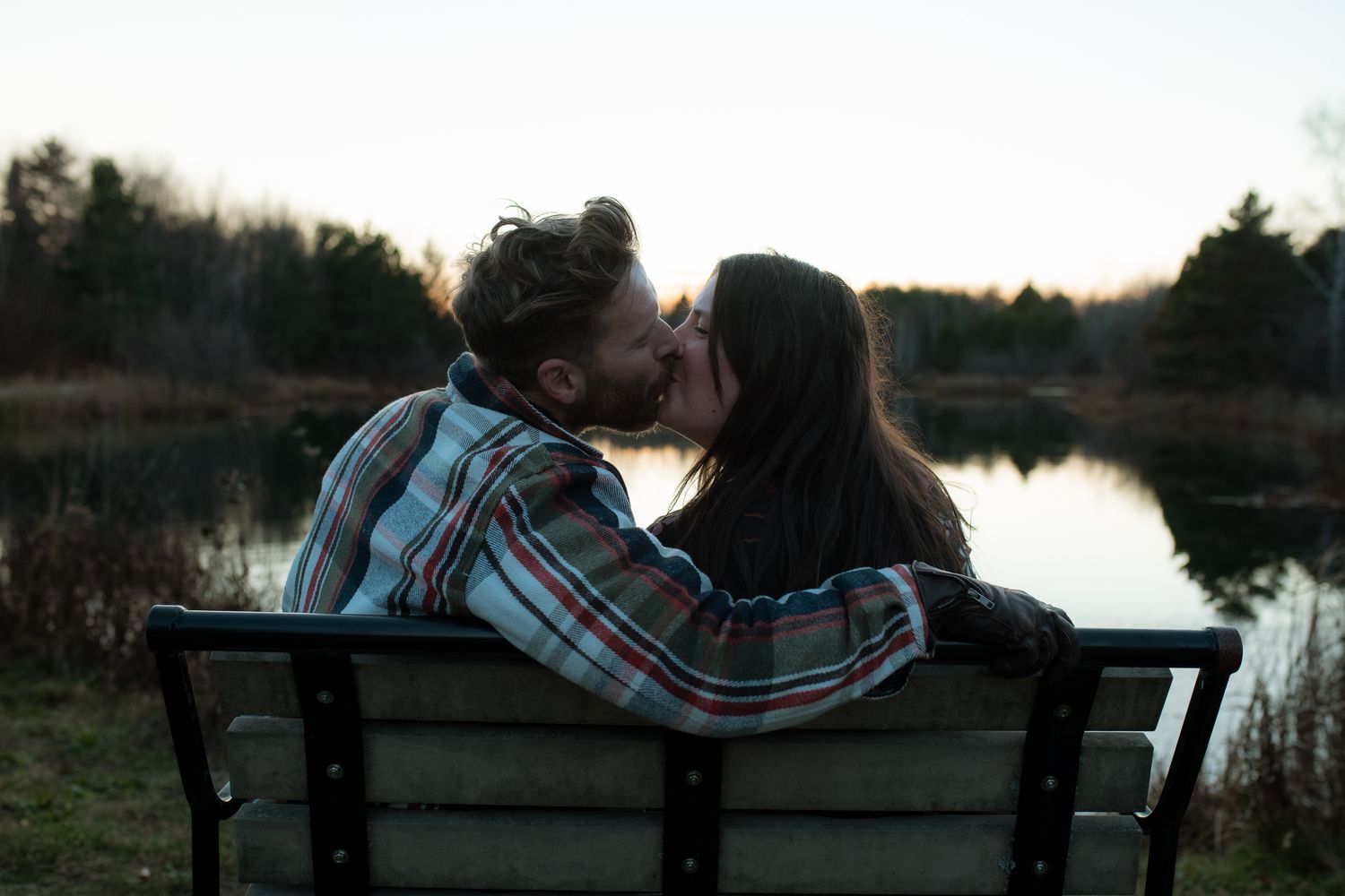 A couple sits together on a bench by a tranquil lake during sunset creating a romantic silhouette.
