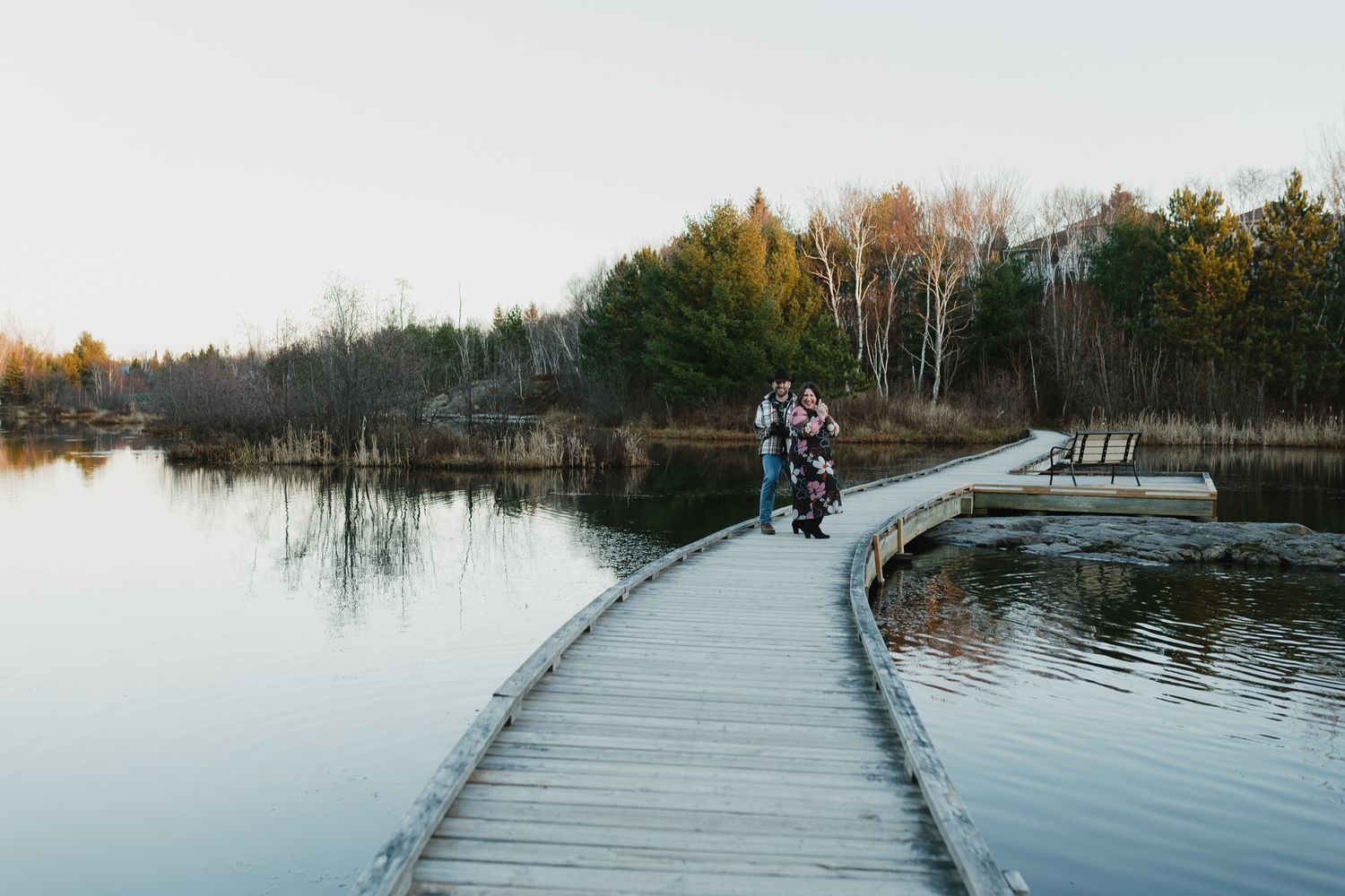 People walking along a wooden boardwalk over calm lake waters surrounded by autumn trees in a peaceful natural setting.