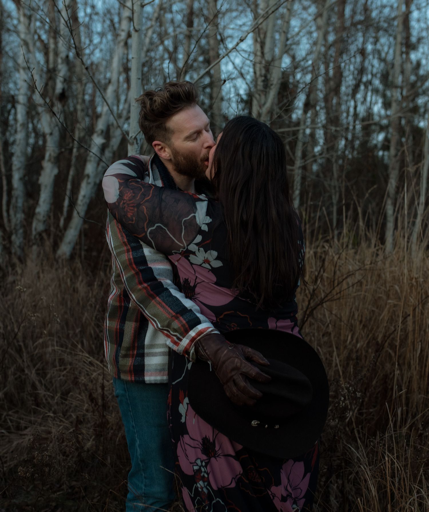 A couple embraces in an intimate moment among bare winter trees and tall grass at dusk.