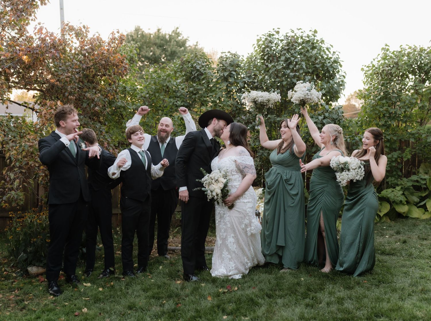 Wedding party celebrating outdoors, bridesmaids in green dresses, groomsmen in black suits, bride and groom kissing.