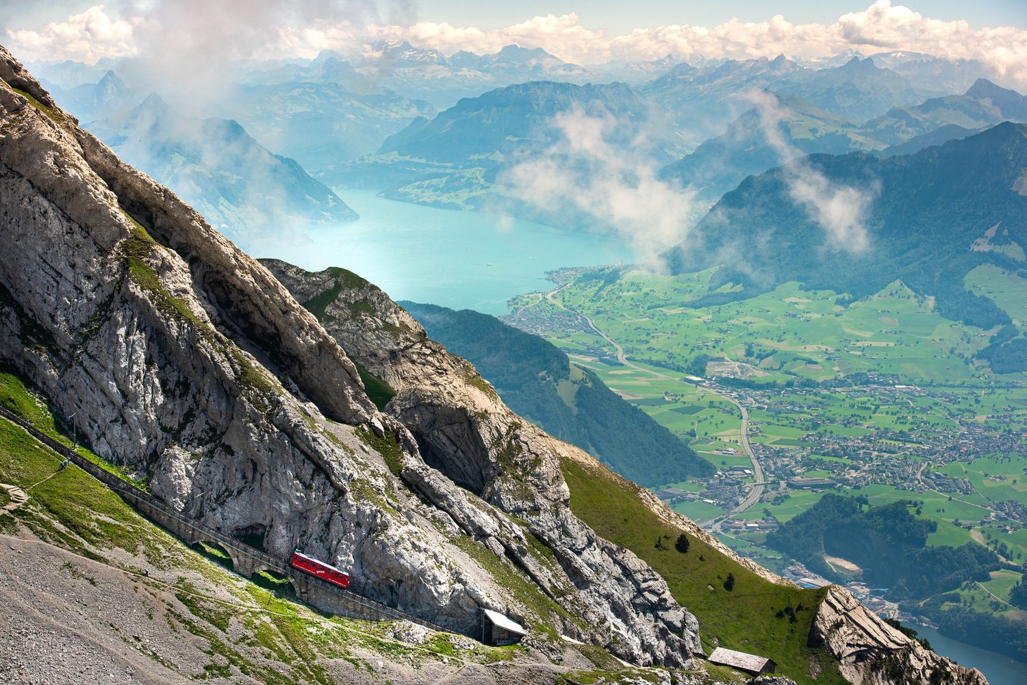 Red cogwheel train on steep Mount Pilatus slope with Lake Lucerne and Swiss Alps valley panorama below.
