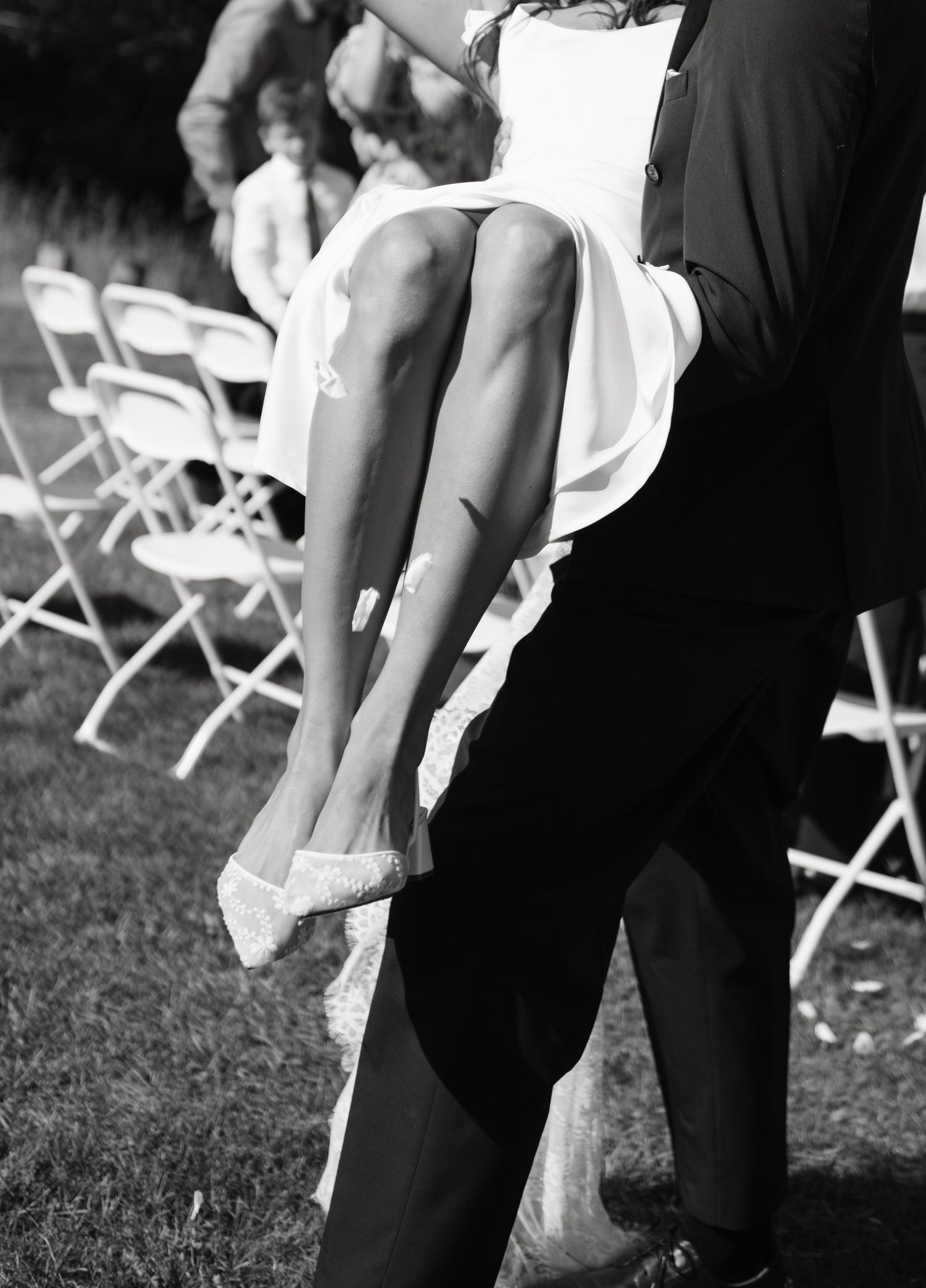 Black and white photo of a bride being carried by a groom at an outdoor wedding ceremony with white chairs.