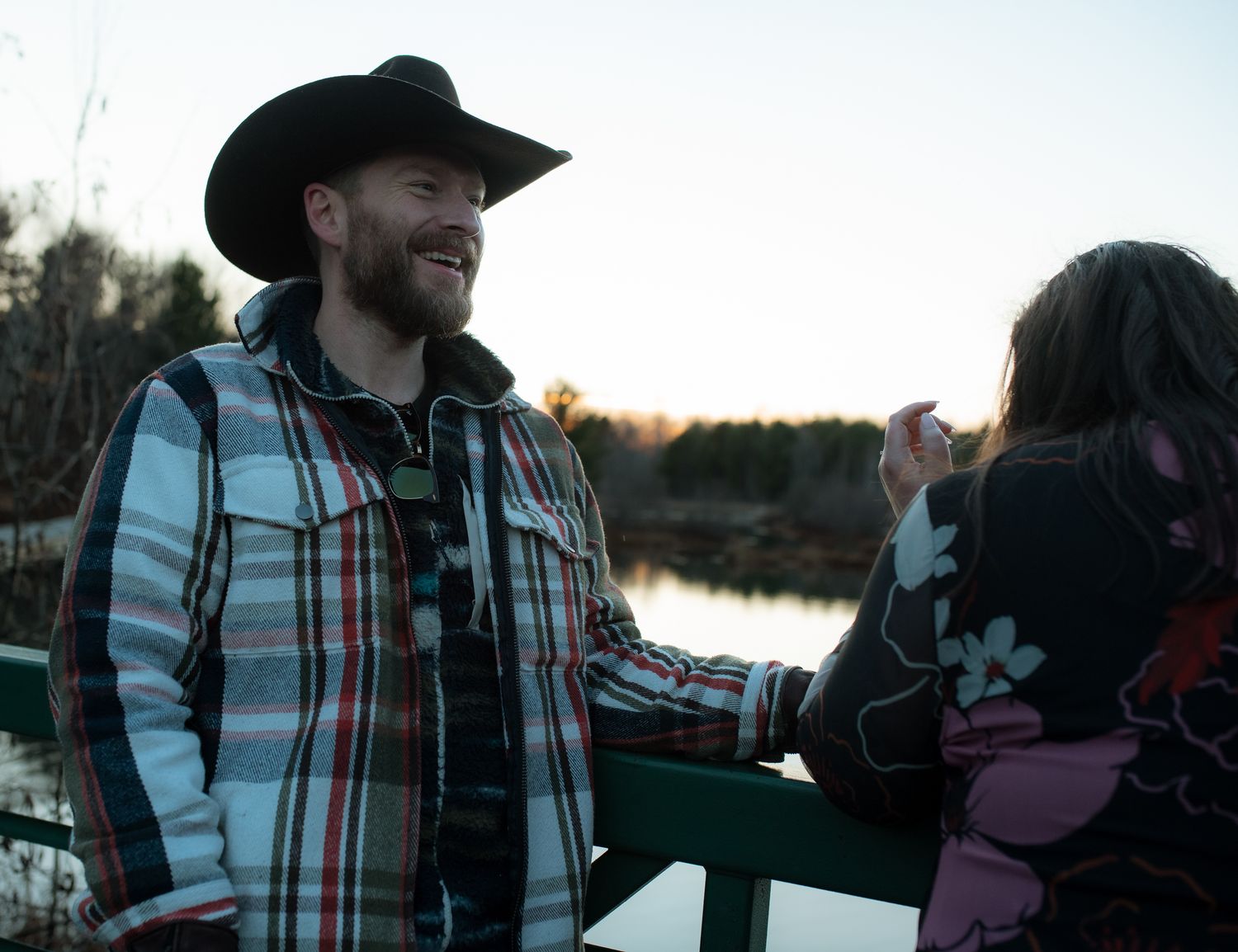A group of people in plaid shirts and hats enjoying an outdoor gathering at sunset near a lake.