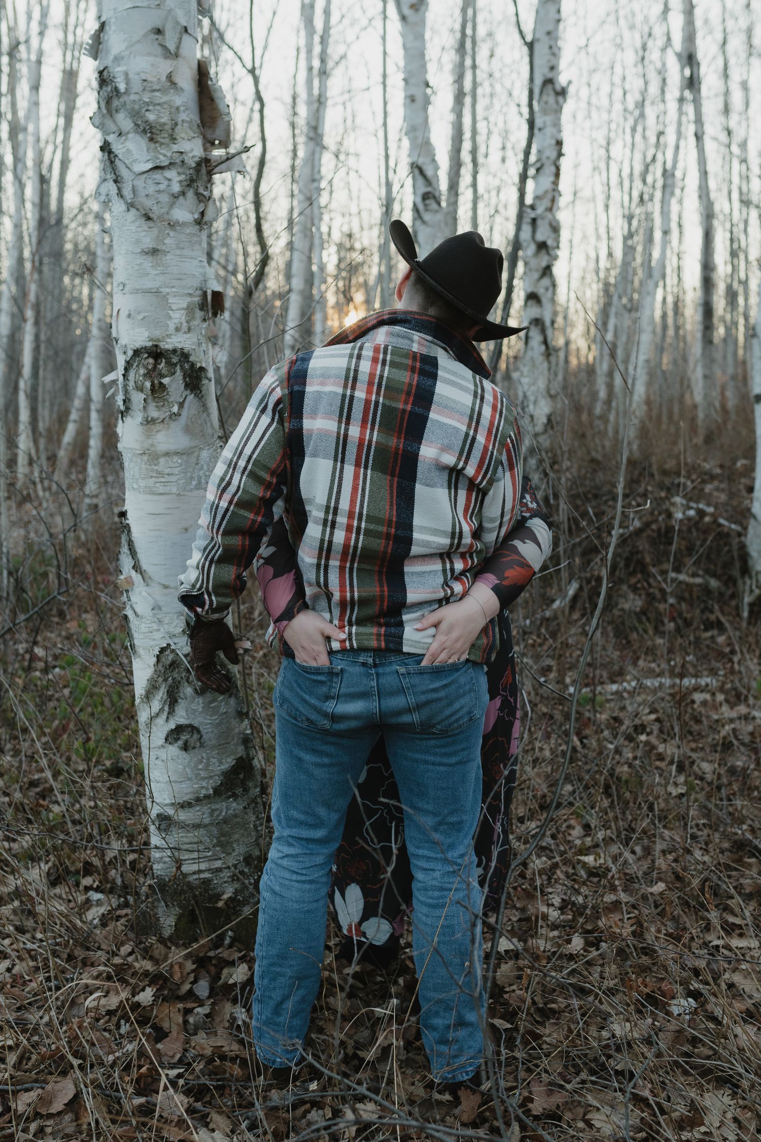 Person in plaid shirt and jeans stands among white birch trees in a winter forest landscape.