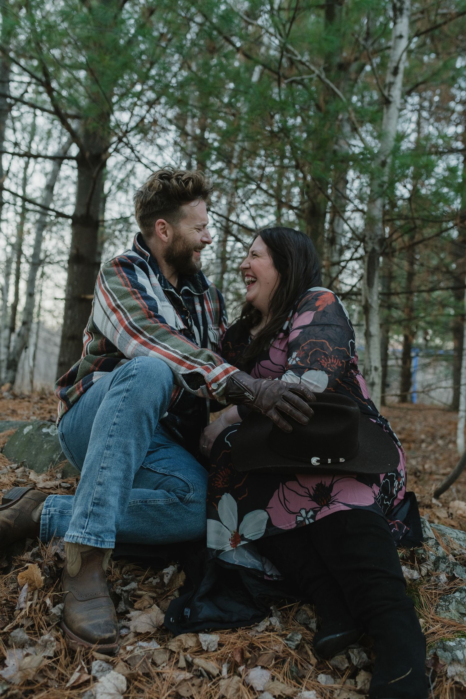 A couple sits together laughing on fallen leaves in a misty forest setting wearing casual fall clothing.