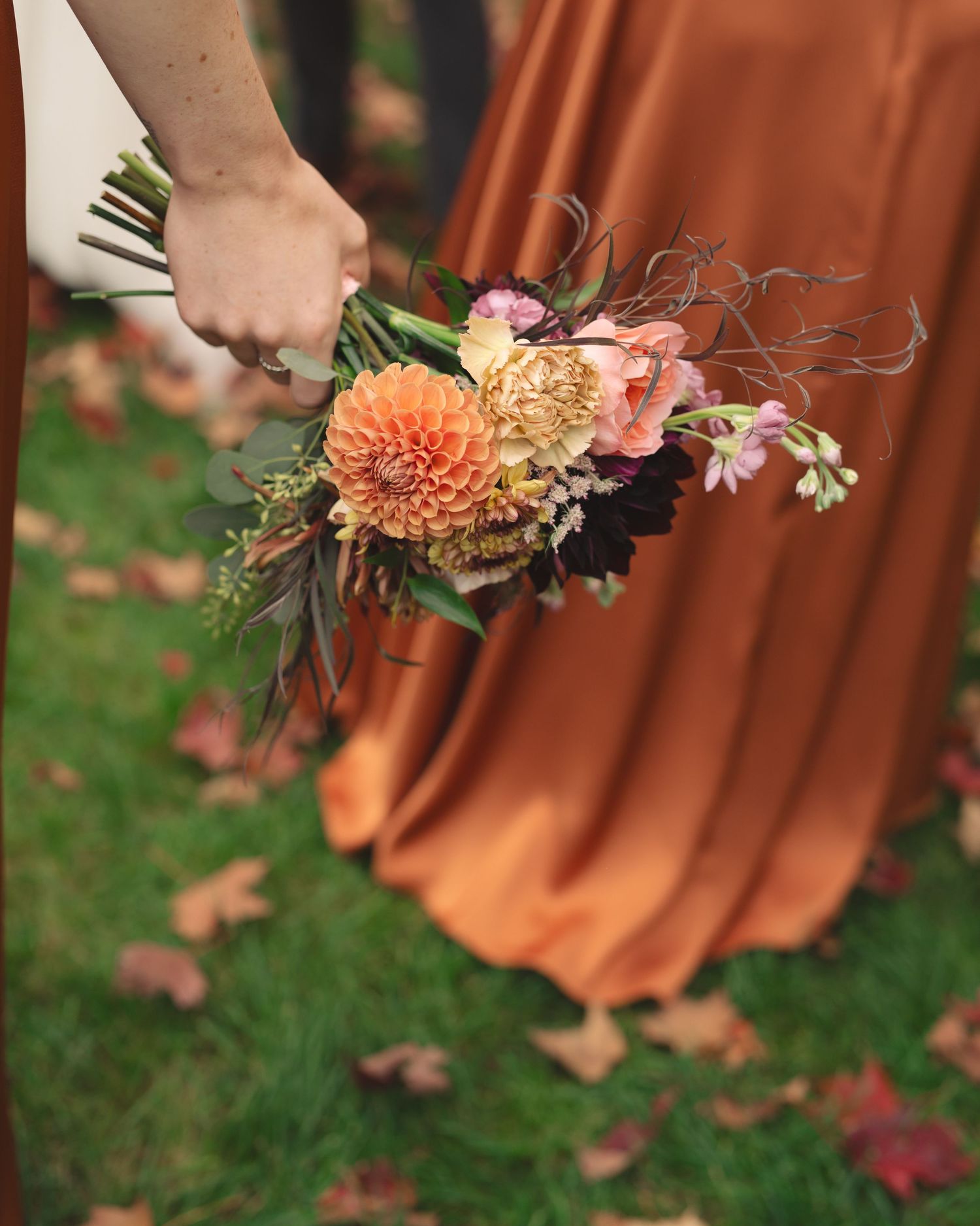 Hand holding an autumn floral bouquet with orange dahlias against a rust-colored dress and fallen leaves on grass.