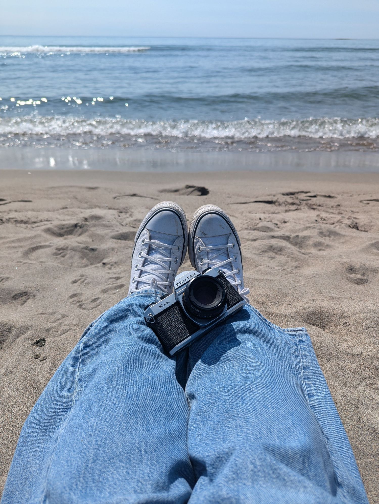 Person relaxing on sandy beach wearing blue jeans and sneakers with ocean waves in background.