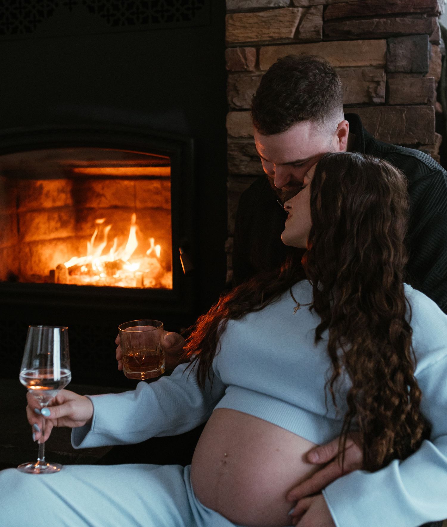 Couple sharing a kiss by a roaring fireplace, pregnant woman holding a wine glass.