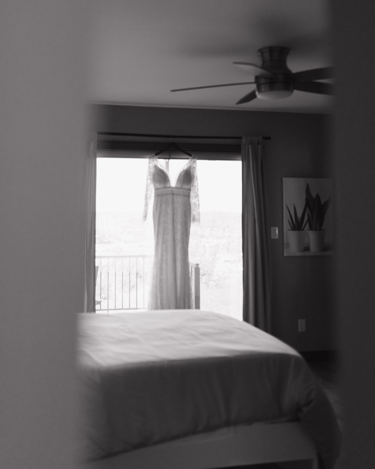 Black and white photo of an elegant wedding dress hanging by a bedroom window with a ceiling fan above a neatly made bed.