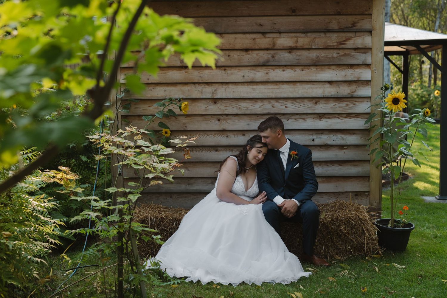 Bride and groom sitting on hay bale beside rustic wooden barn with sunflowers, sharing an intimate moment outdoors.