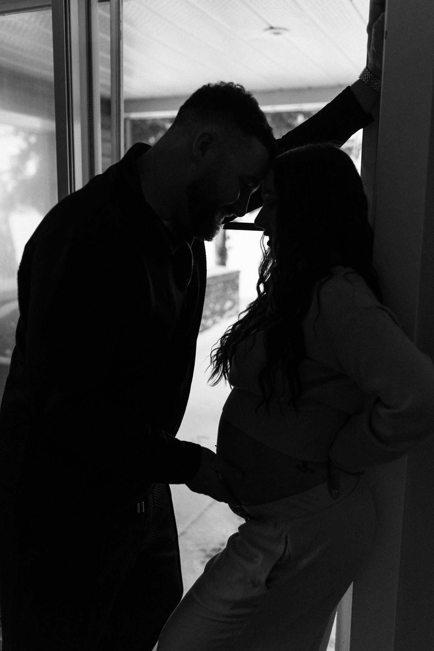 Black and white silhouette of a couple holding hands and leaning close together near a bright window.