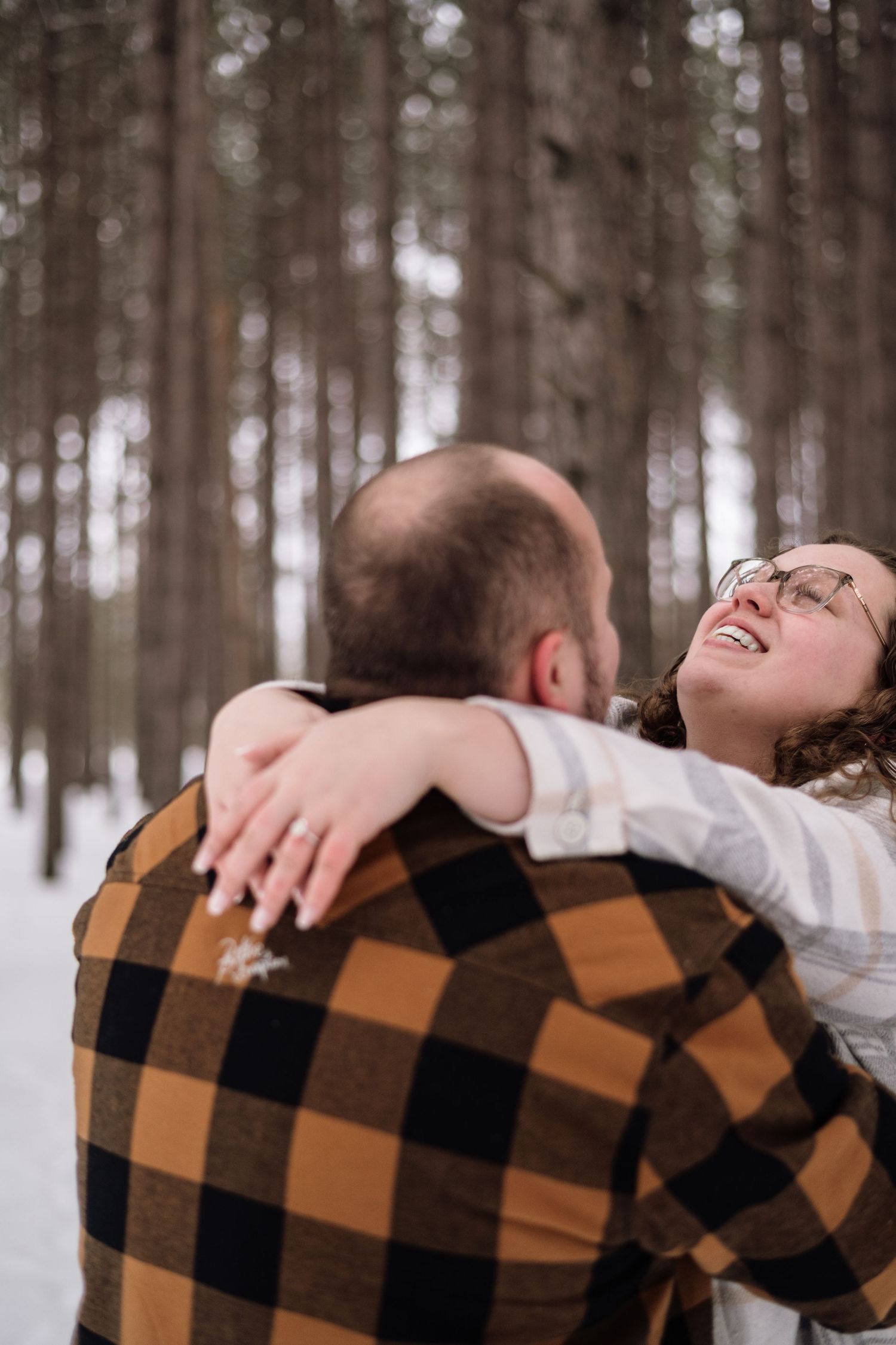Couple embracing and laughing in snowy winter forest, woman wearing glasses and engagement ring hugs man in plaid flannel shirt.