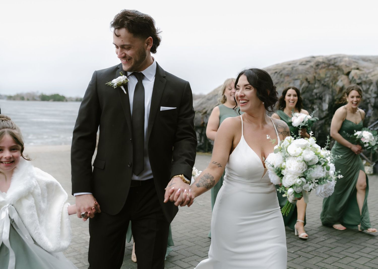 Bride in white gown and groom in black suit walk joyfully with bridesmaids in green dresses at waterfront wedding.