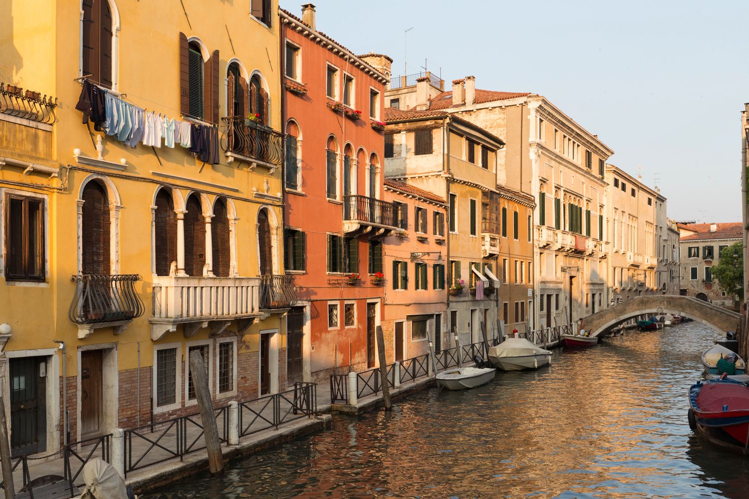 Colorful historic buildings lining a serene Venice canal with moored boats and a stone bridge at golden hour.