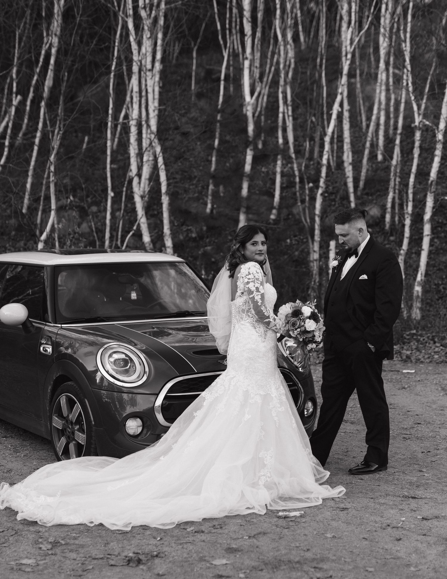 A wedding couple poses with their Mini Cooper car surrounded by birch trees in a black and white photo at Laurentian Conservation in Sudbury