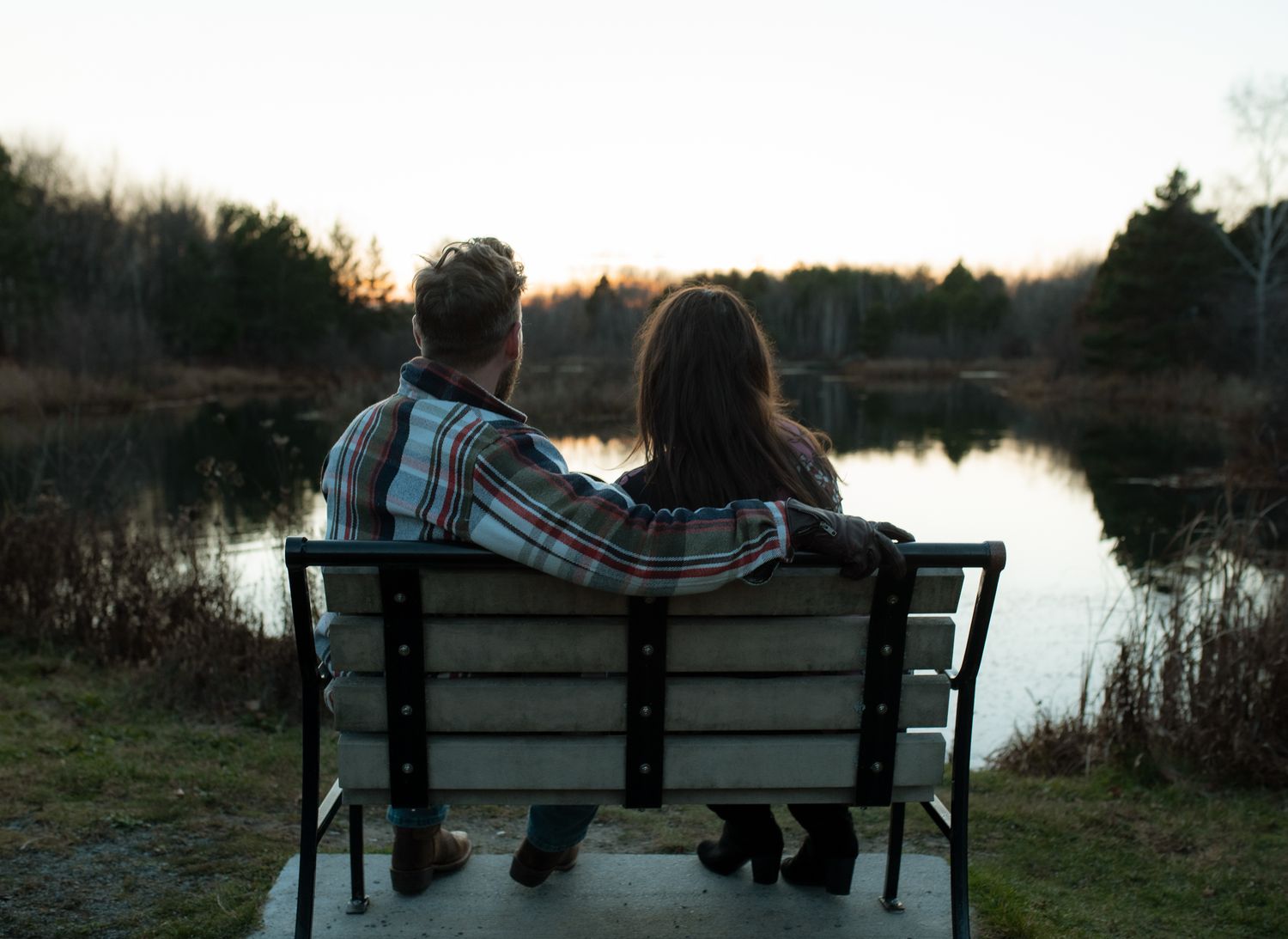 Silhouetted figures sit on a park bench overlooking a tranquil lake at sunset.