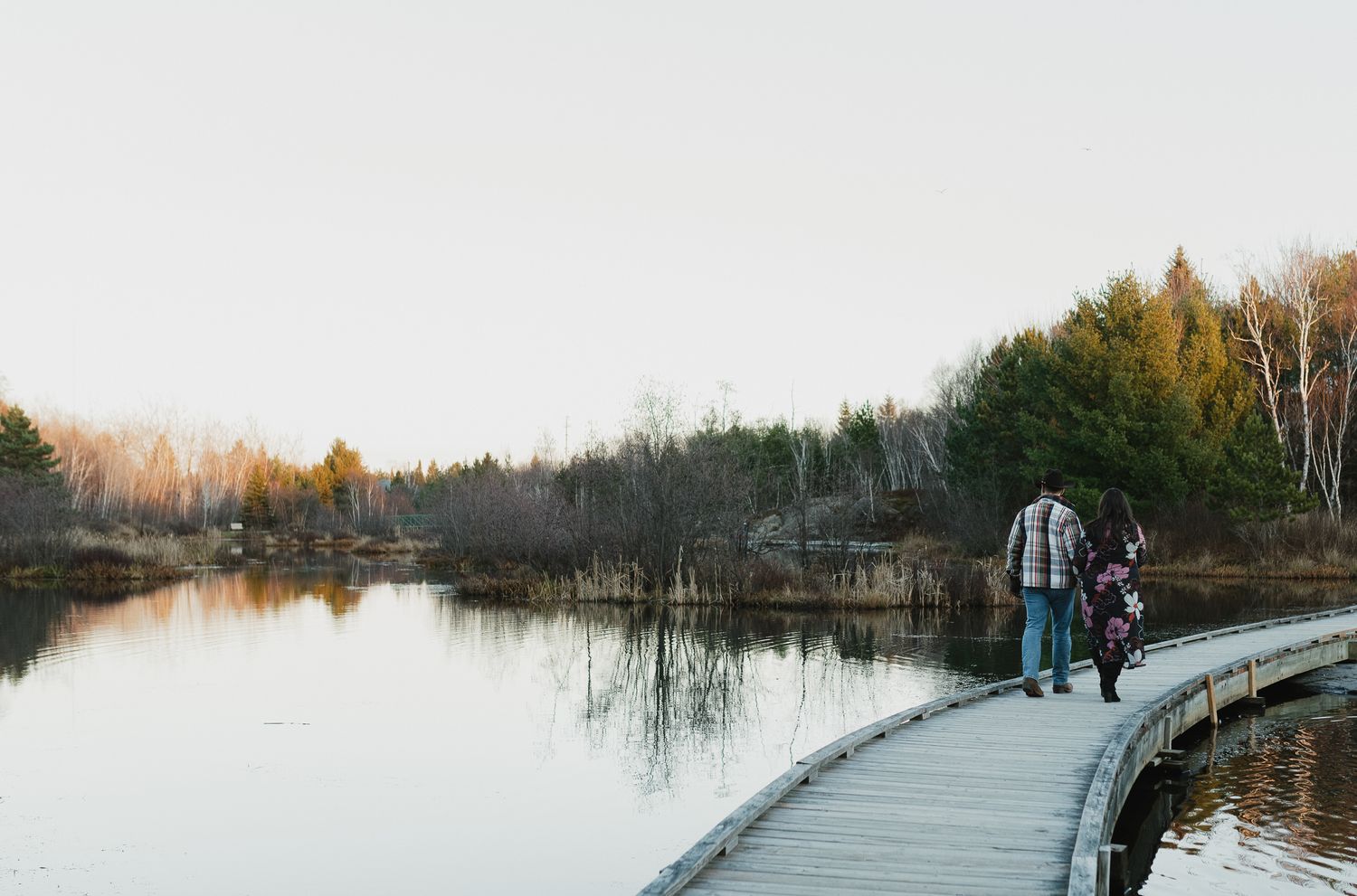 People walk along a curved wooden boardwalk over a misty lake during autumn with bare trees in the background.