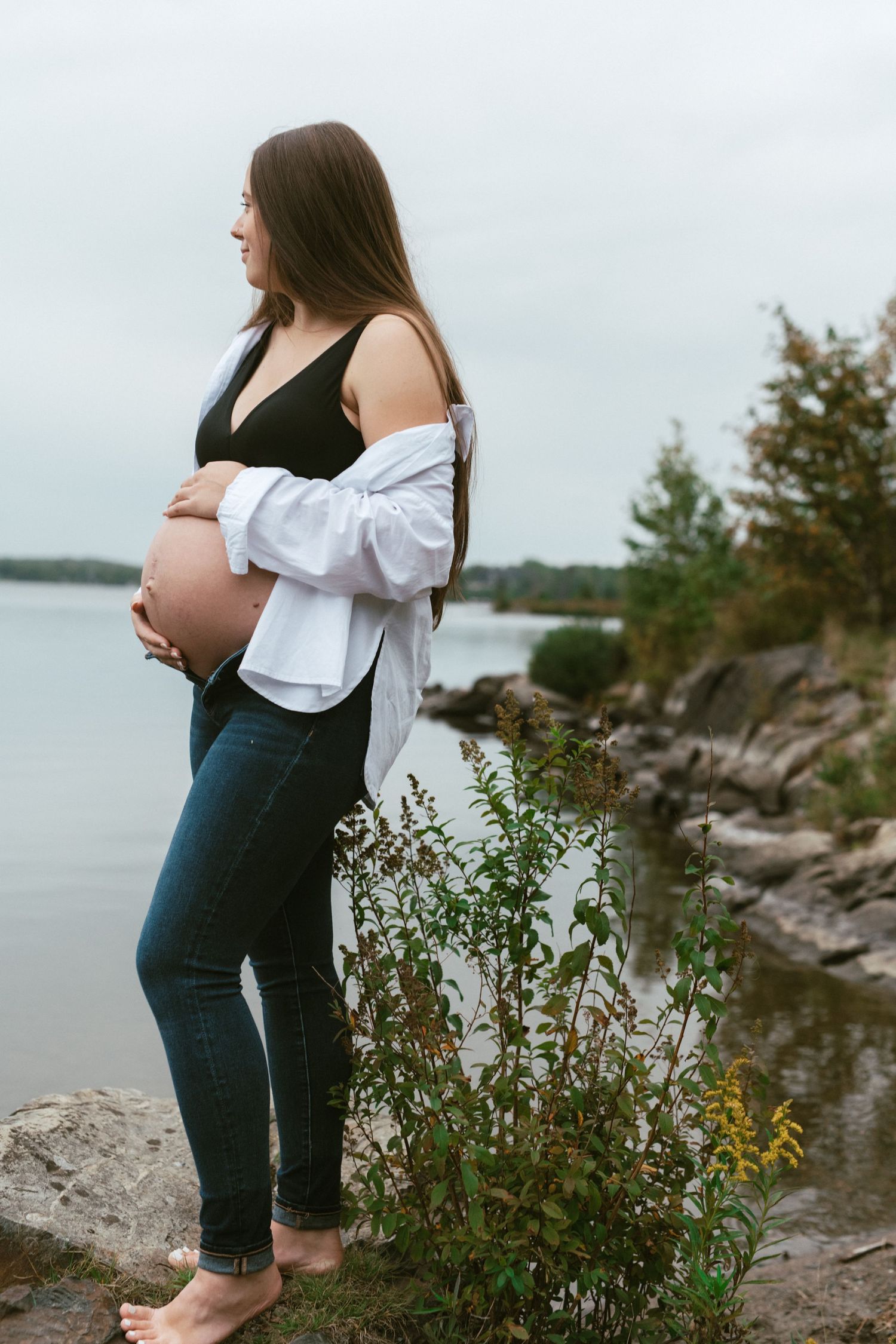 A woman in jeans and white top stands at the rocky shoreline of a lake on an overcast day.