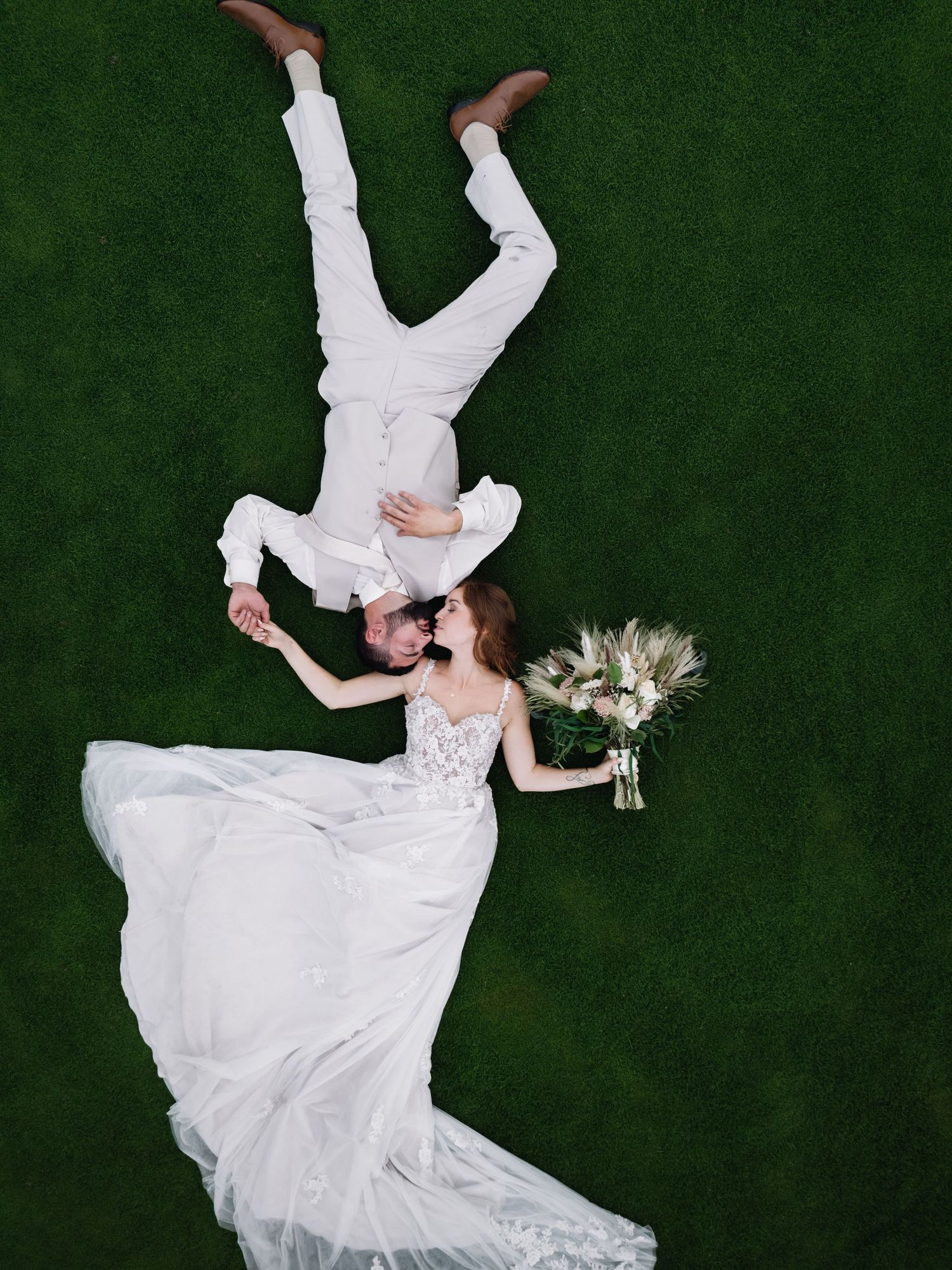 A couple in wedding attire lying on green grass with white floral bouquet in artistic overhead shot.