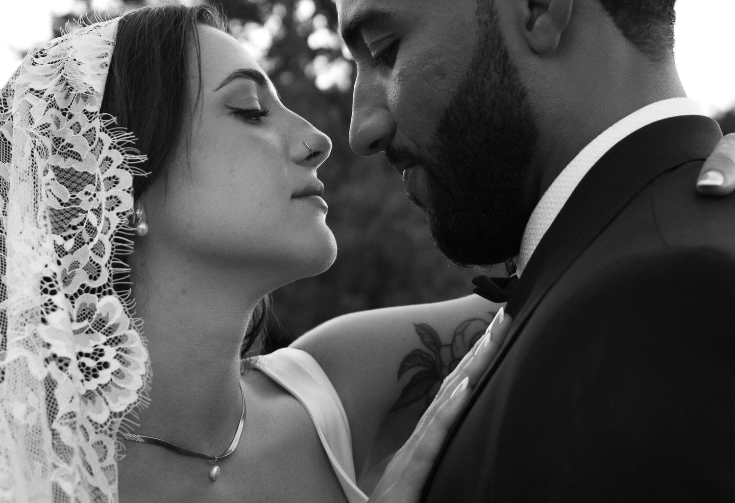 Bride in lace veil and groom in tuxedo share an intimate close moment in black and white wedding photo.