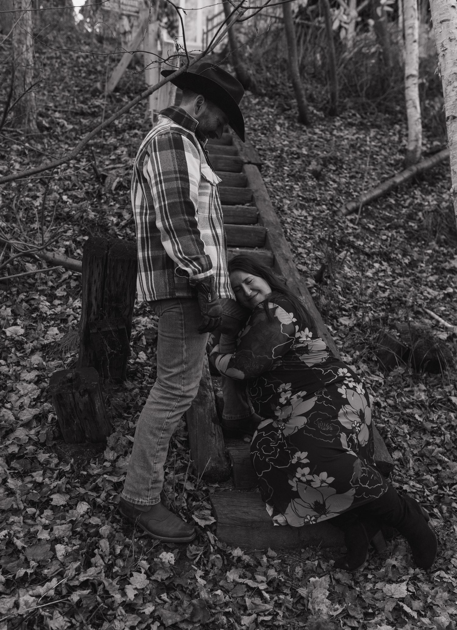 A black and white photograph shows a person in plaid and denim standing on rustic wooden steps in a wooded area.