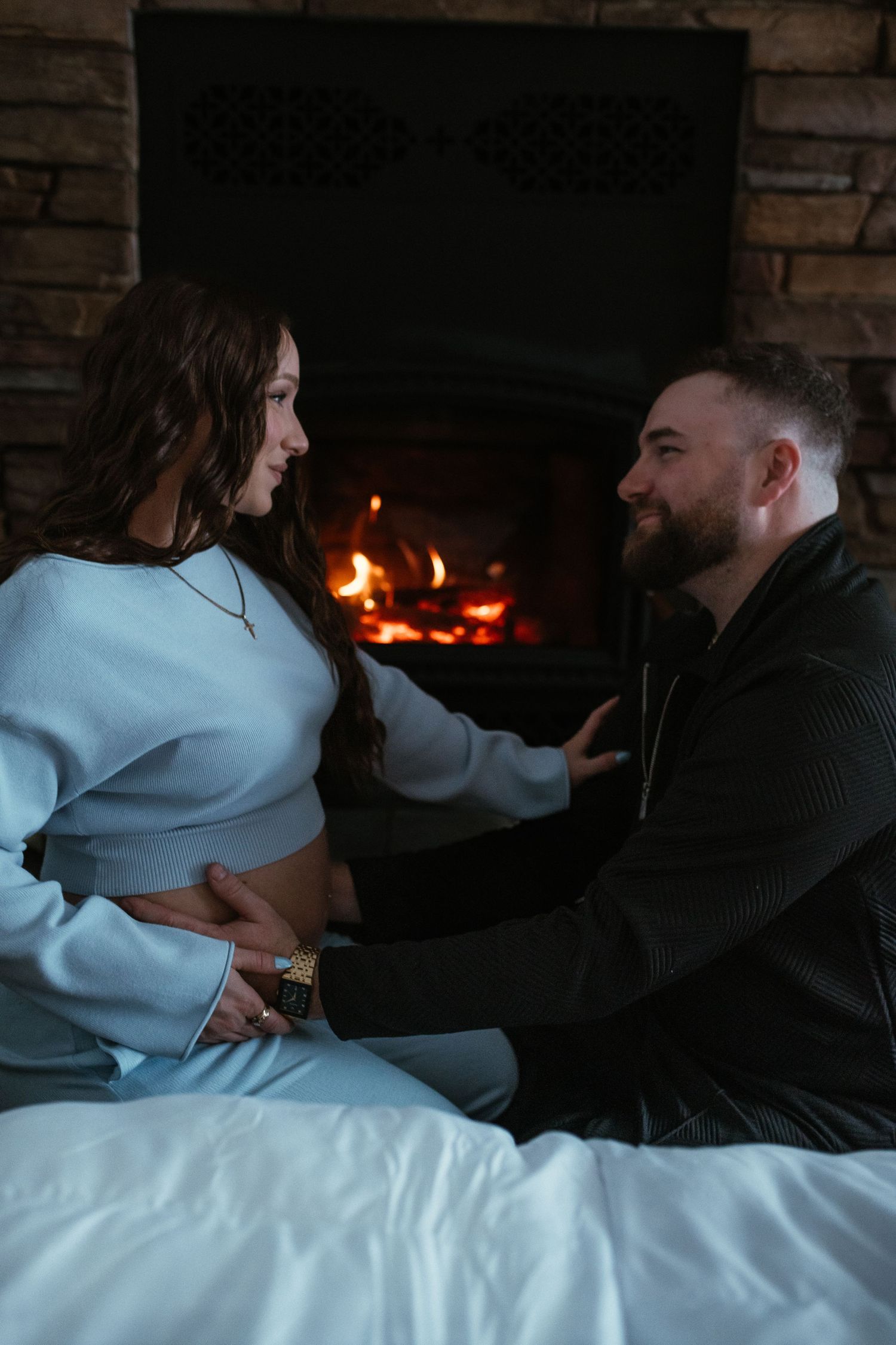 Smiling couple sharing a tender moment by fireplace, man touching pregnant womans belly.