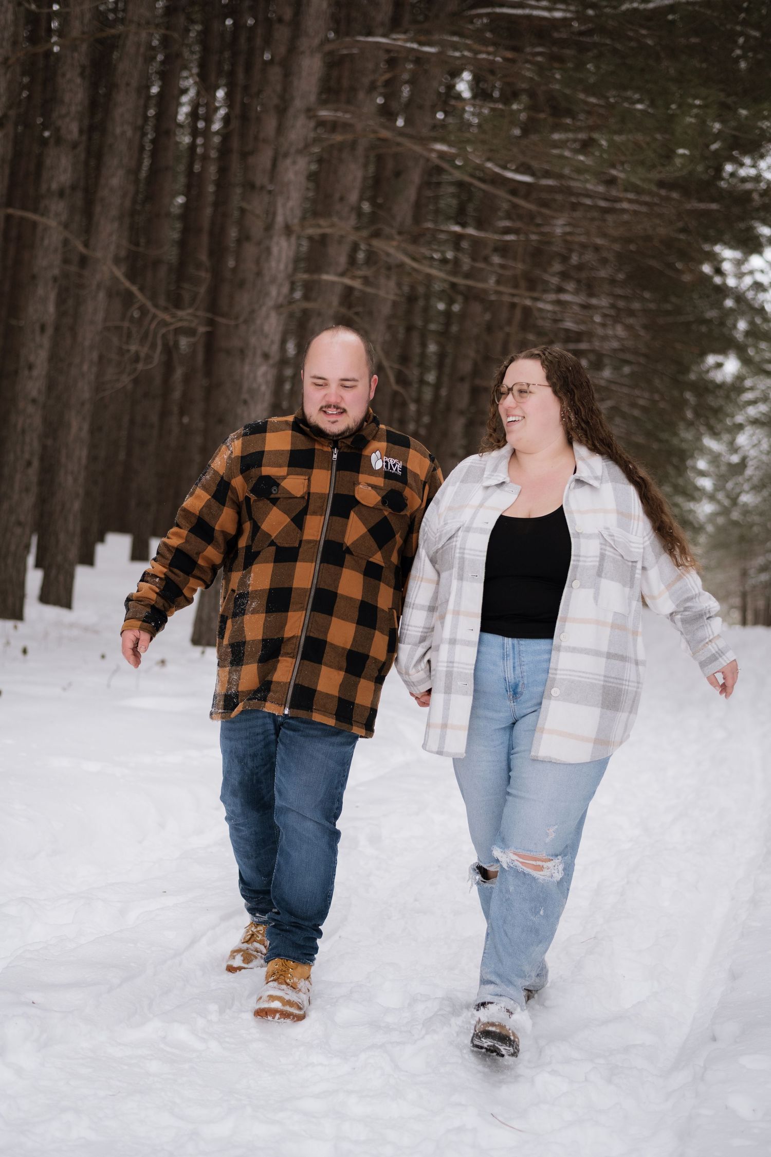 Couple holding hands walking on snowy forest path, both wearing plaid shirts and jeans.