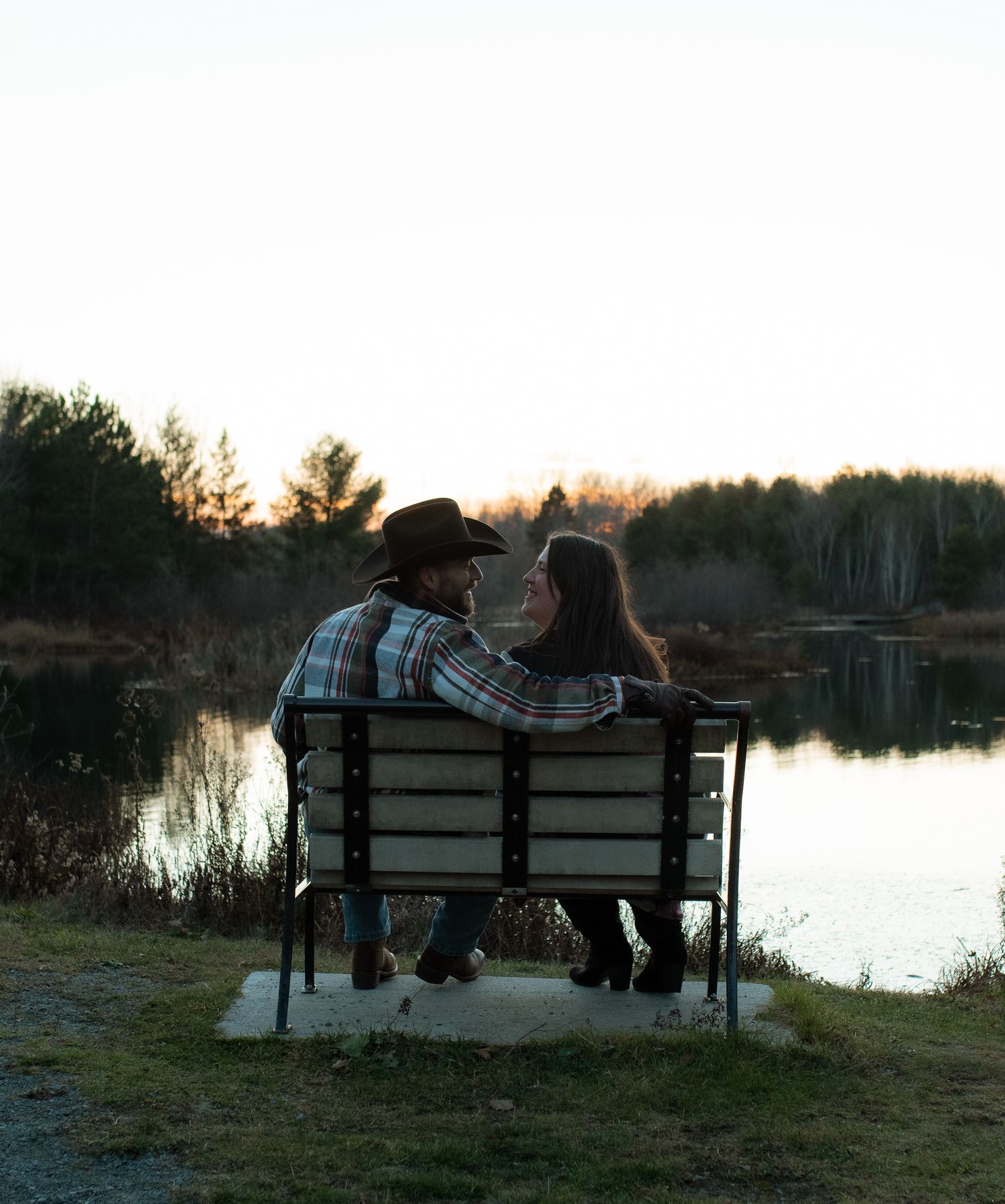 Couple sitting together on wooden bench by tranquil lake at sunset with trees silhouetted in background.