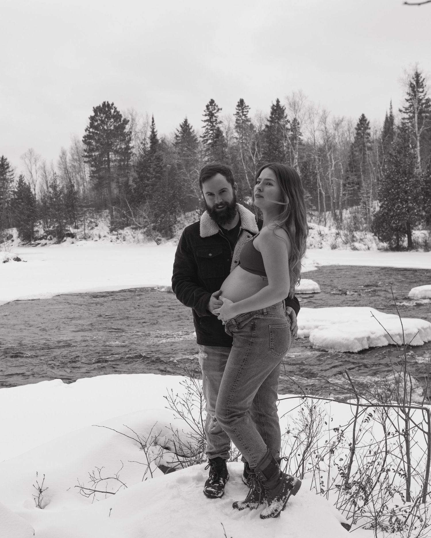 A couple poses together in a snowy winter landscape with evergreen trees in the background.
