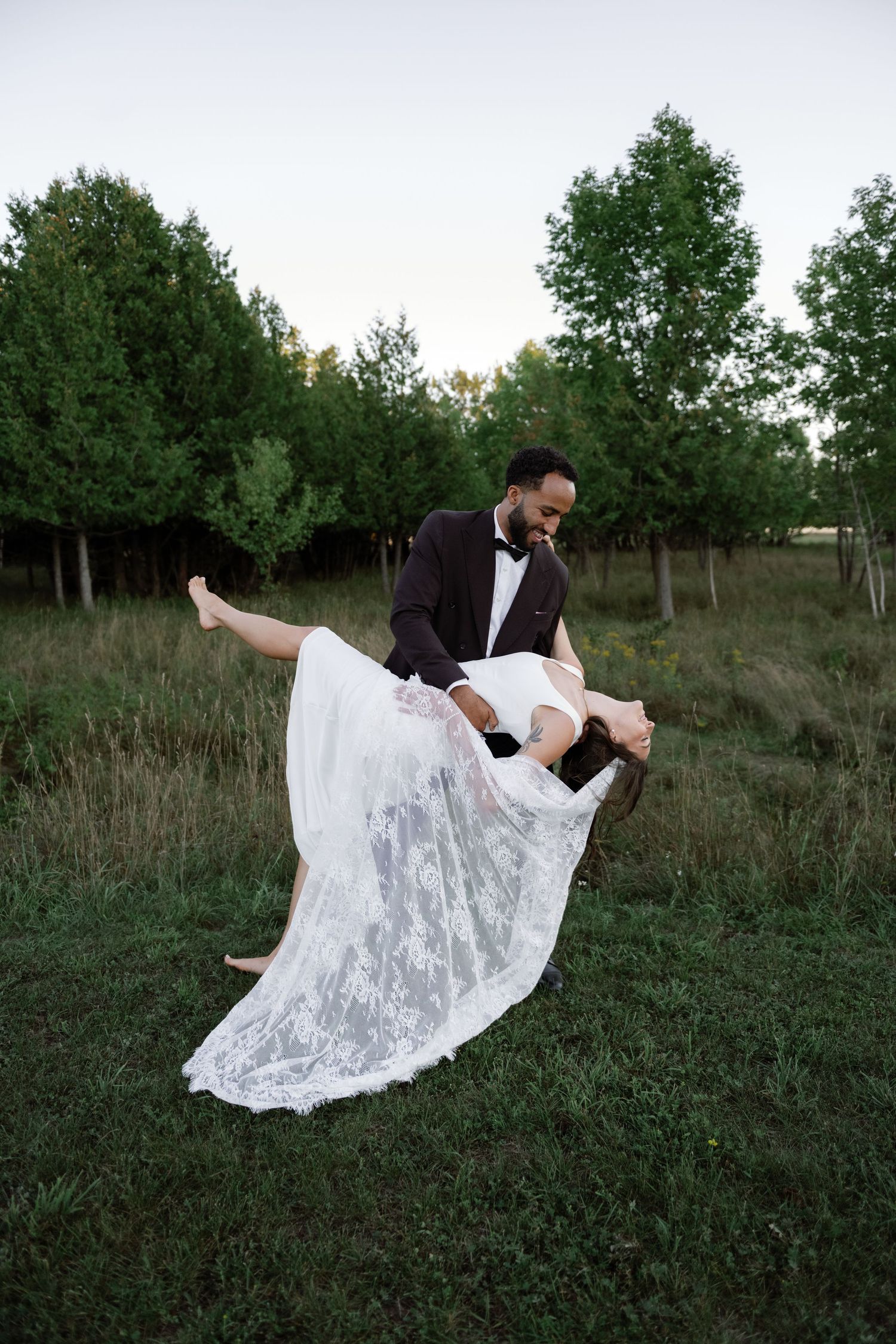 Groom in tuxedo dipping bride in lace wedding gown in a lush green outdoor setting at dusk.