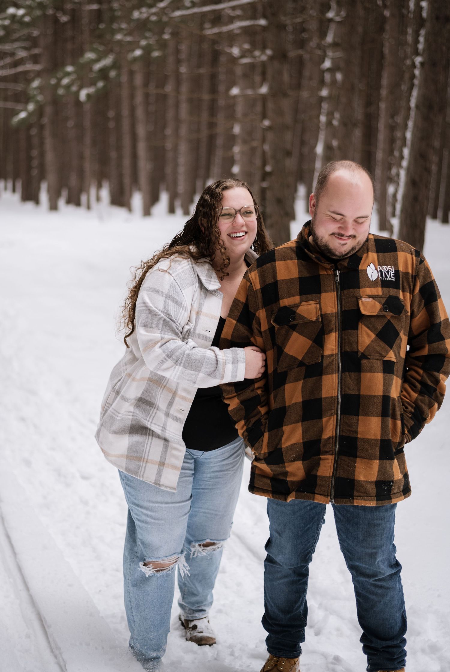 Couple walking together on a snowy forest path in plaid flannel shirts, smiling during a winter engagement photo session.