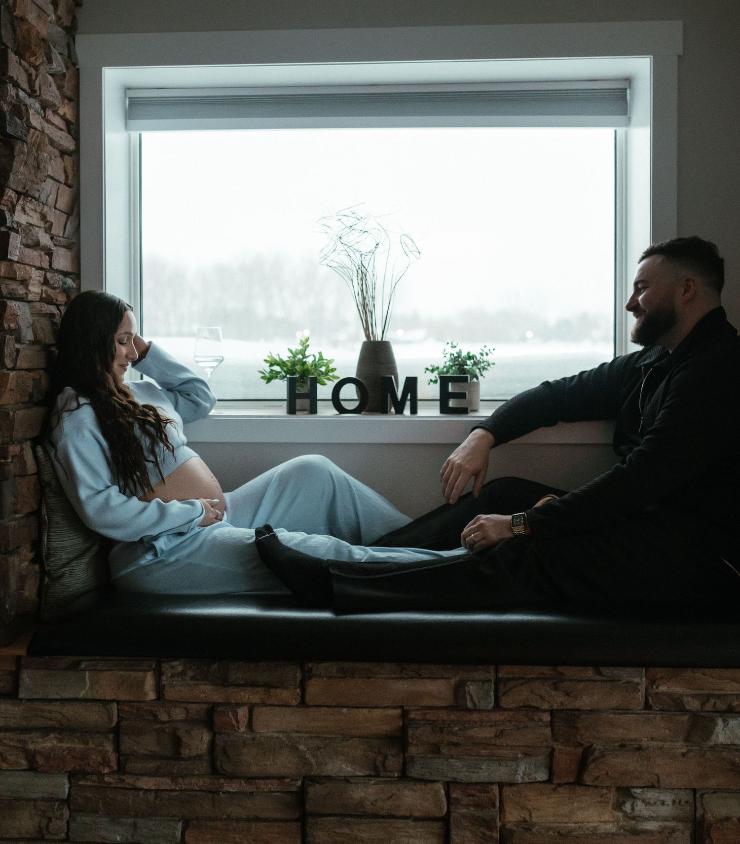 Expecting couple relaxes on stone window seat with HOME sign, sharing a quiet intimate maternity portrait moment.