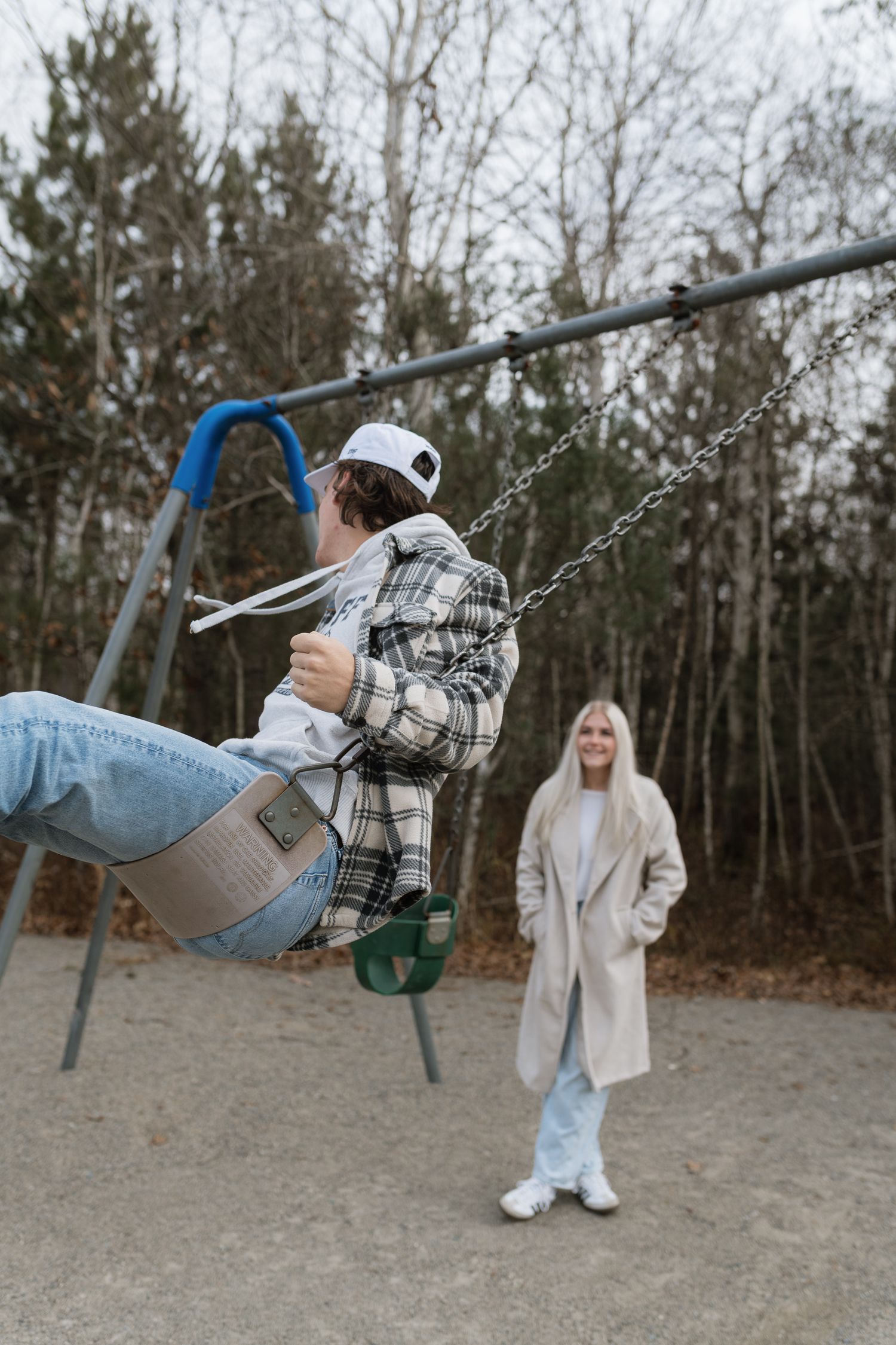 Young man swinging at playground while smiling blonde woman watches in beige coat, autumn park setting.