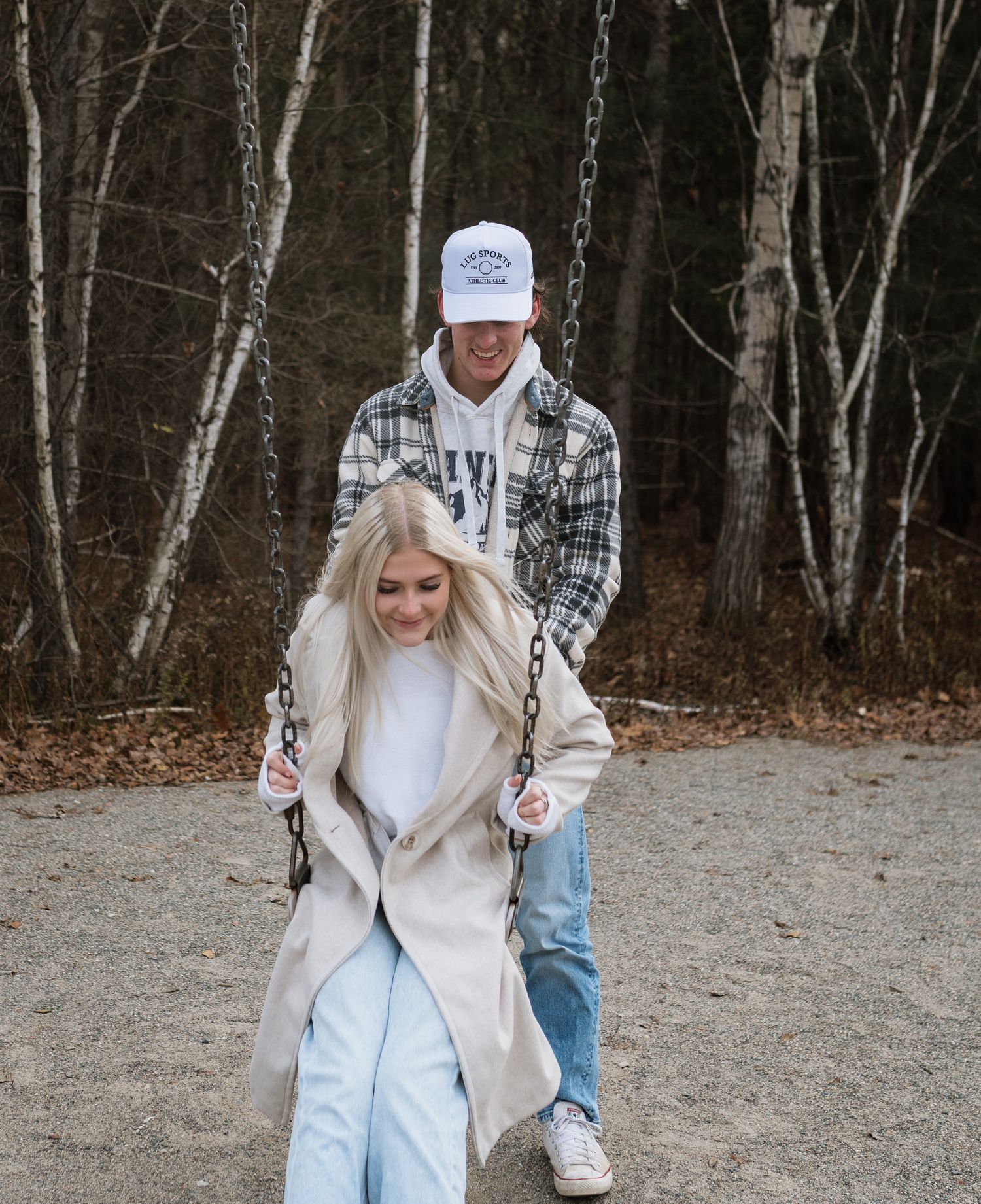 Young man in plaid shirt pushing blonde woman on swing in autumn park setting.