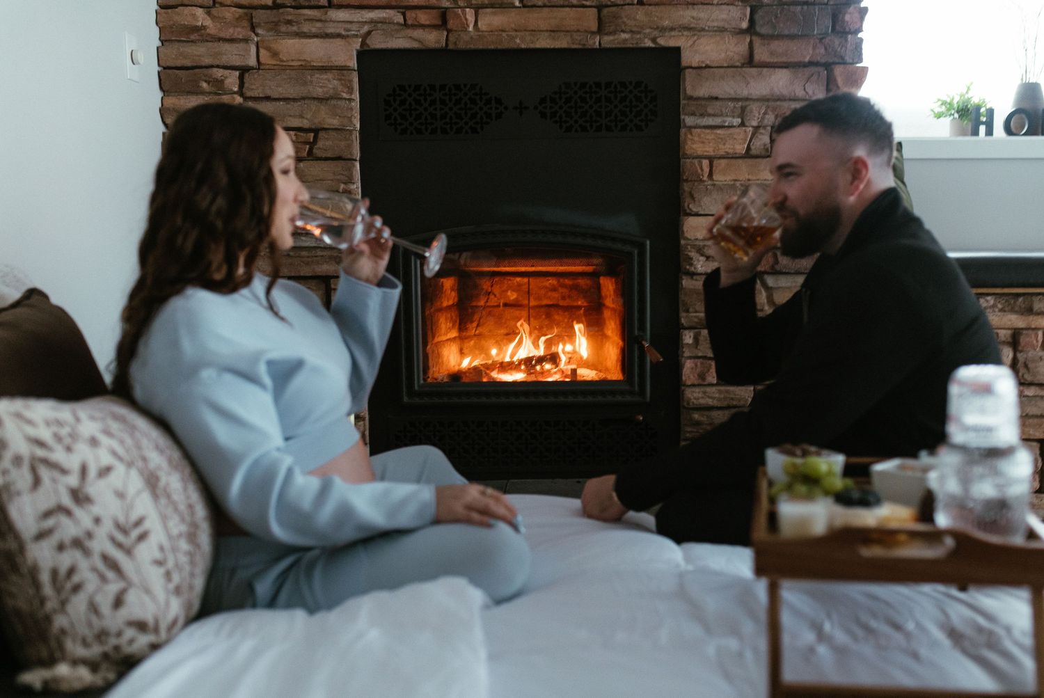 Couple sitting by a cozy fireplace, sipping drinks on a bed in a rustic stone-walled room.