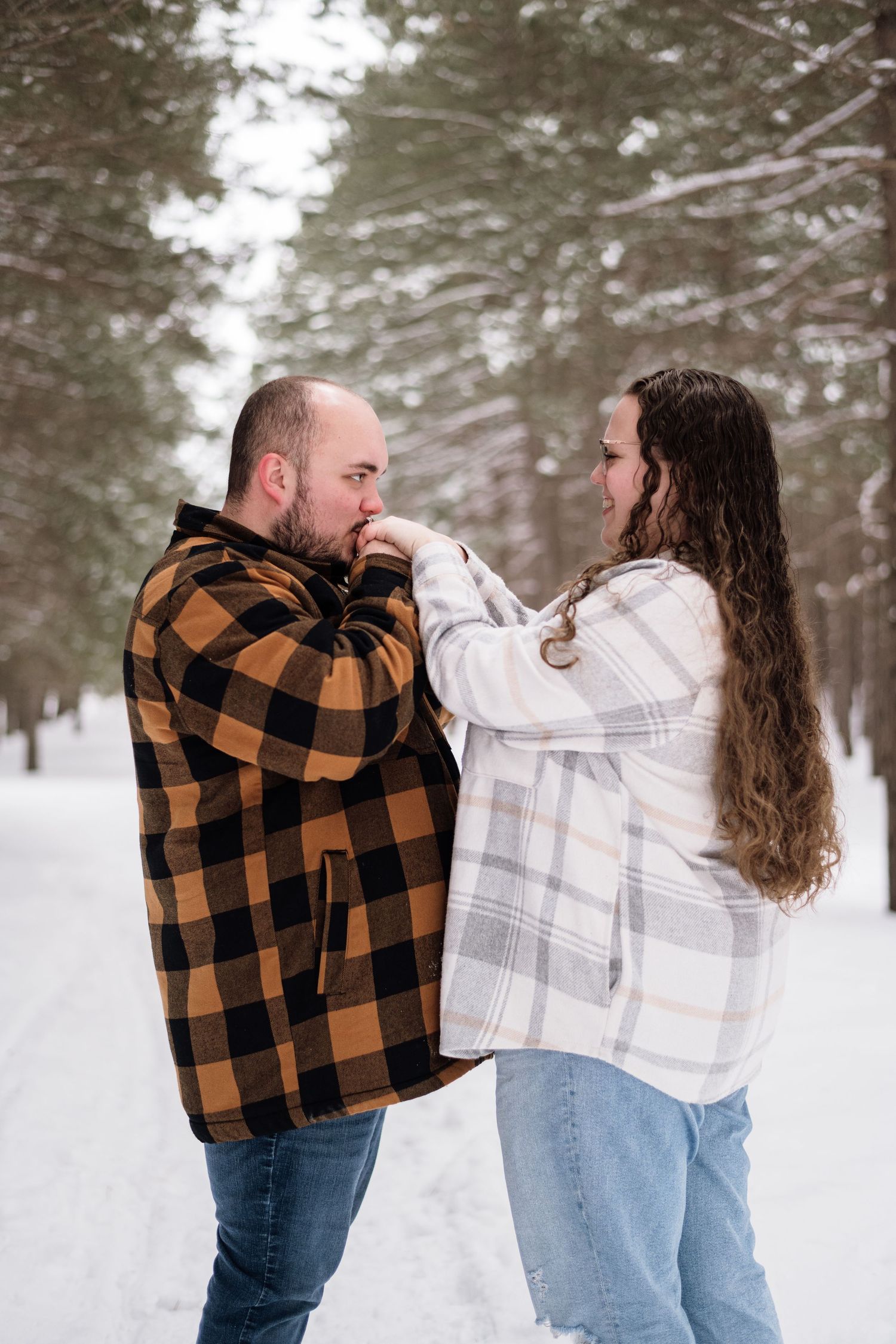 Man kisses woman's hand tenderly as she holds his face in a romantic snowy pine forest scene.