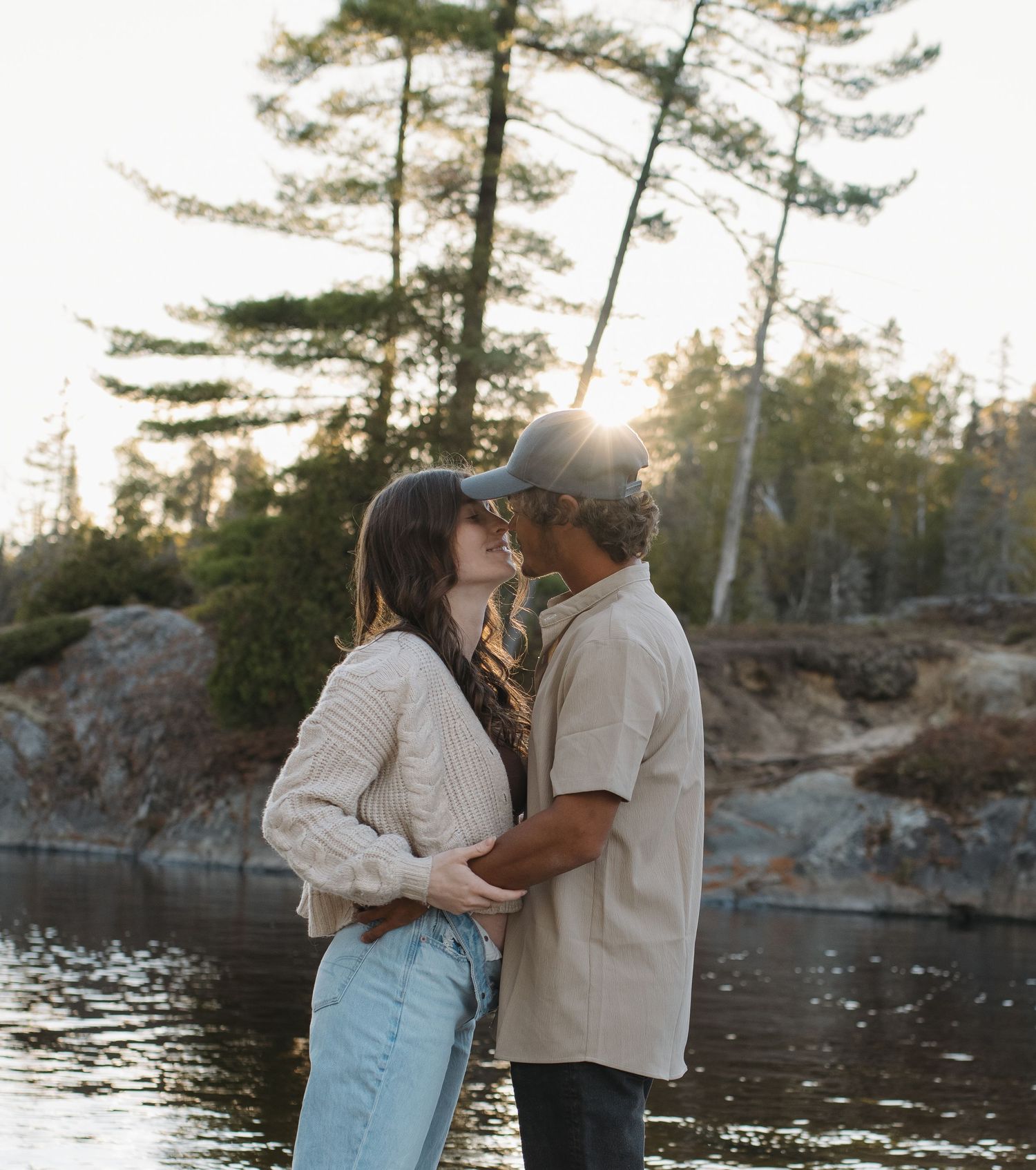 A romantic moment shared between two people on a beach with pine trees and water in the background.