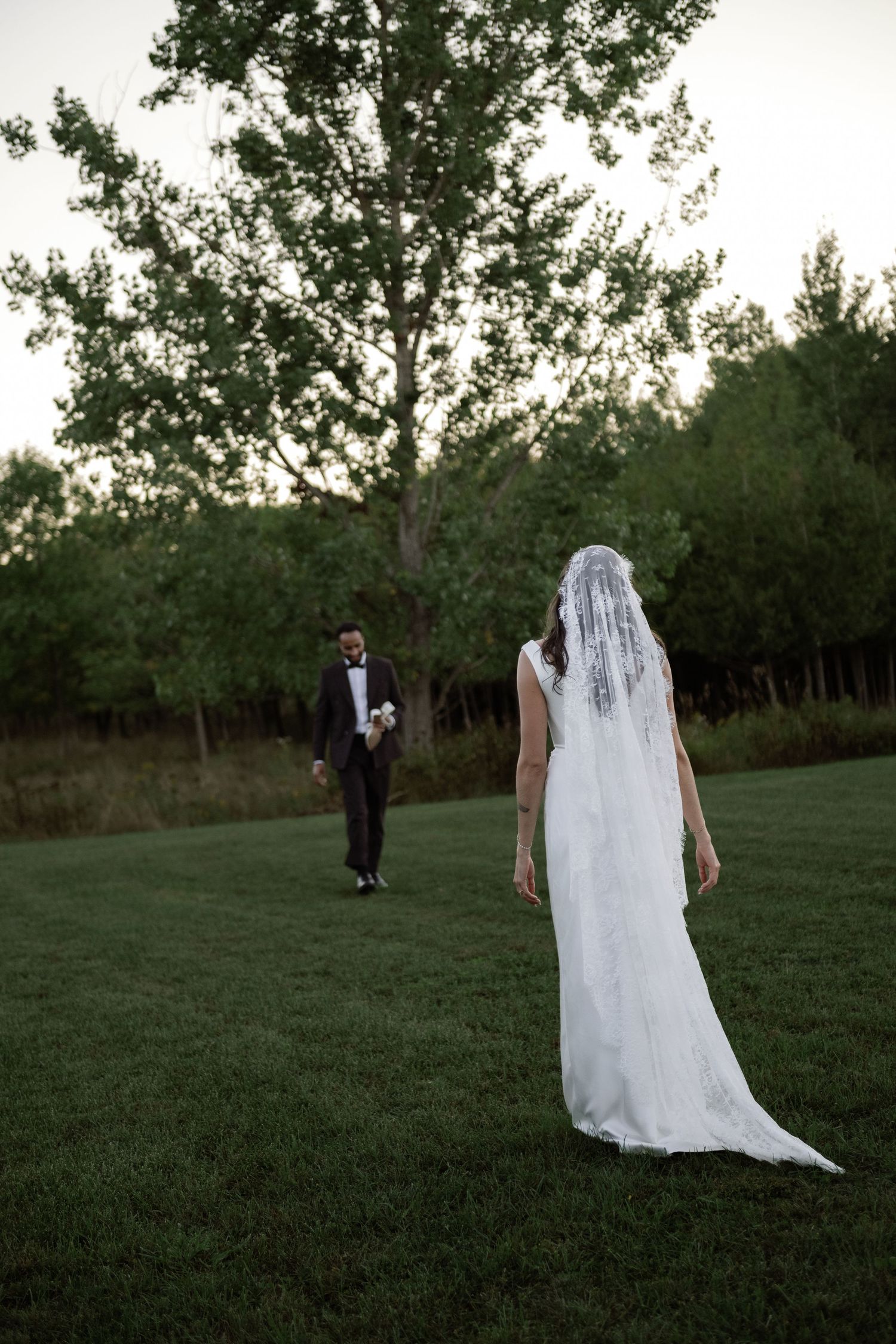 Bride in white lace veil gown and groom in tuxedo standing apart on a green lawn at dusk.