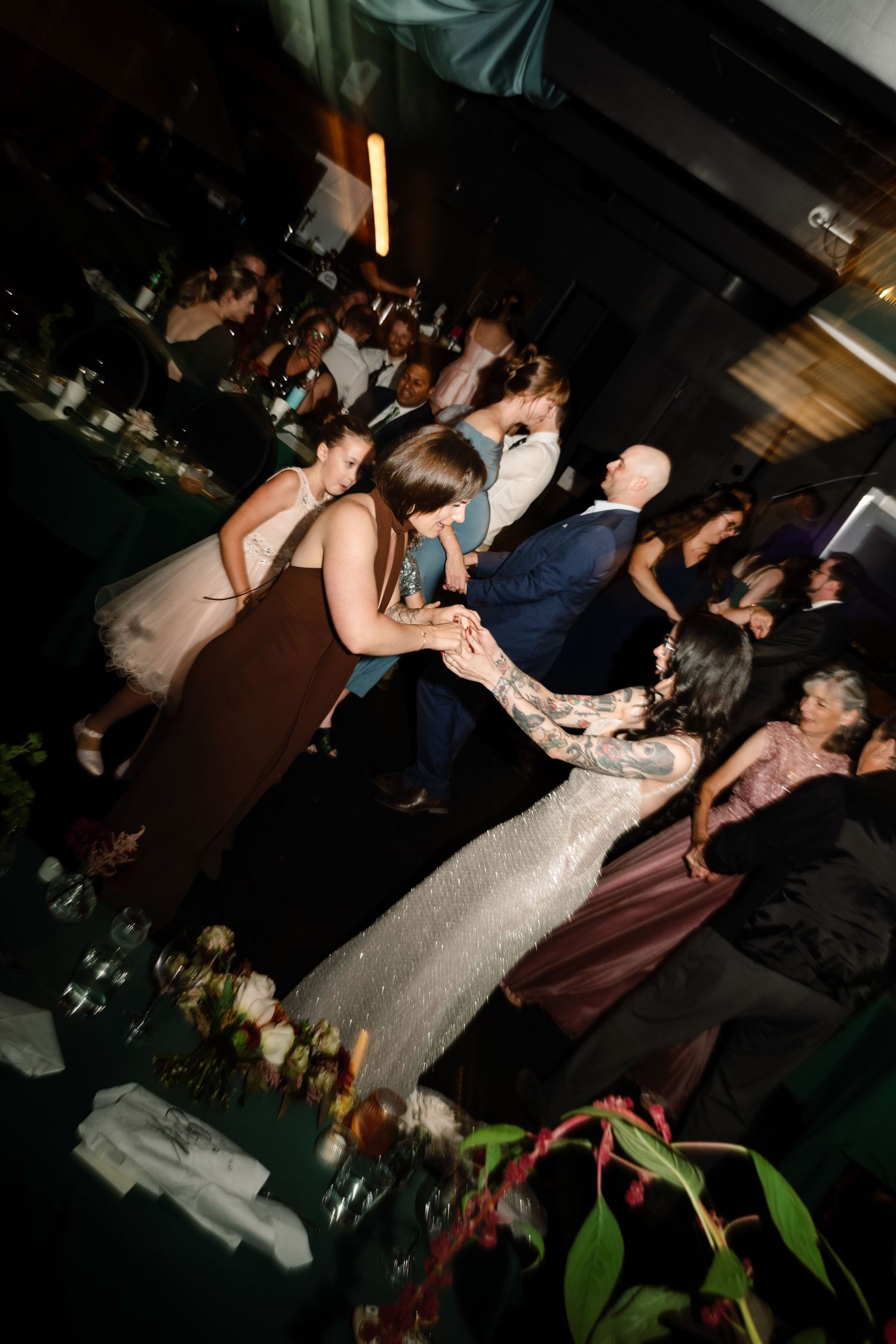 Guests dancing together at an elegant wedding reception with floral centerpieces and dim atmospheric lighting.