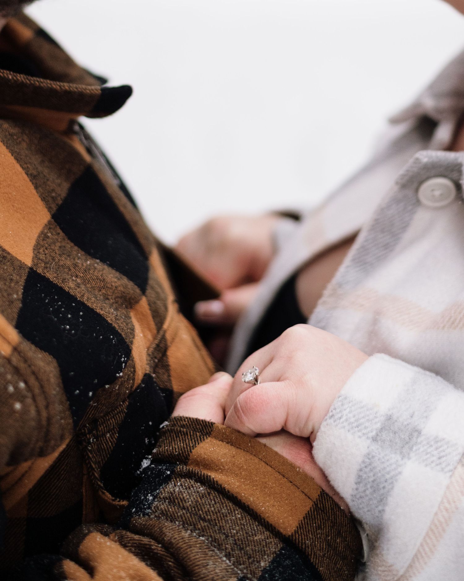 Couple holding hands in winter, woman wearing diamond engagement ring, dressed in plaid flannel jackets outdoors.