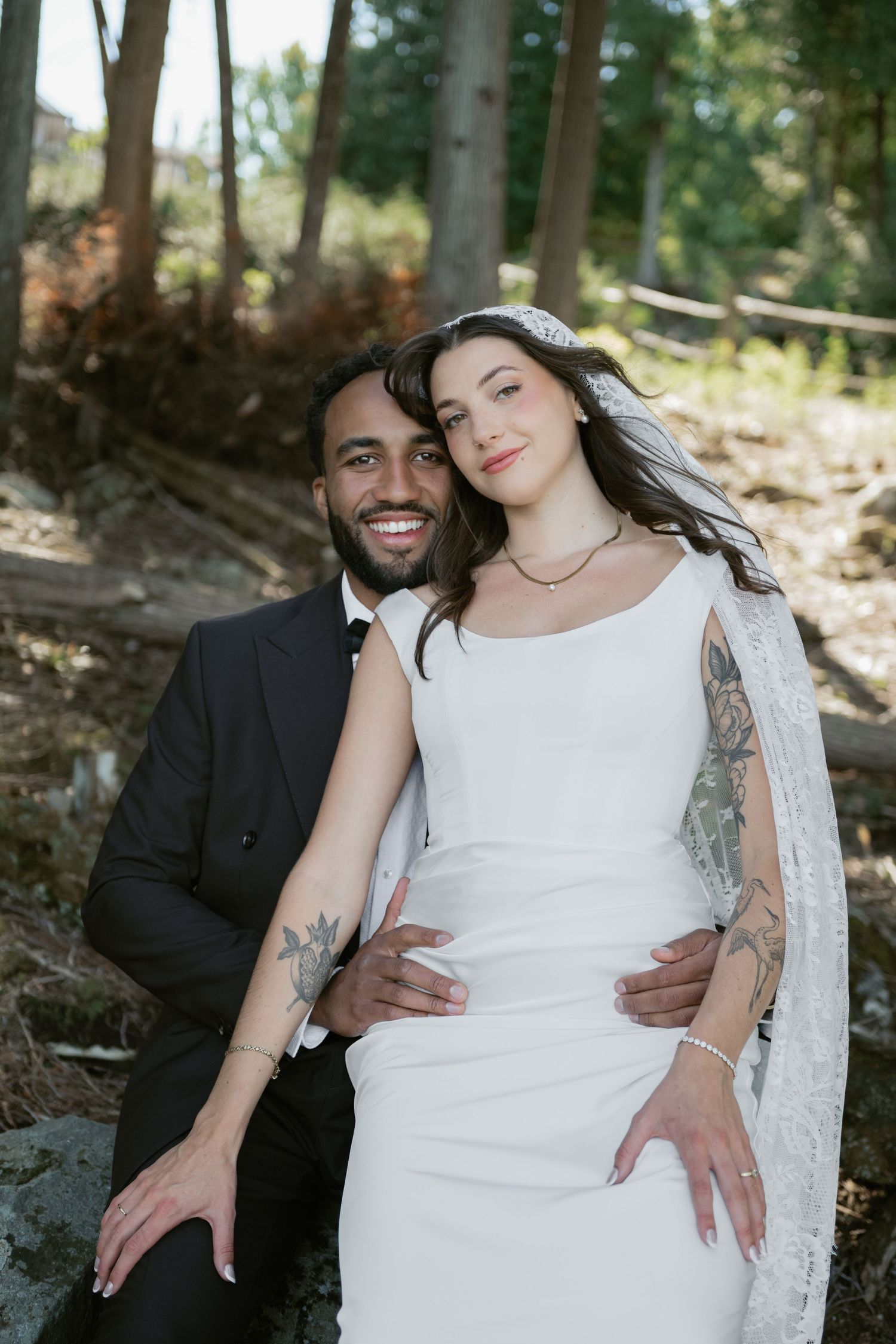 A smiling interracial couple in wedding attire poses outdoors in a forest setting, bride in white gown with lace cape.