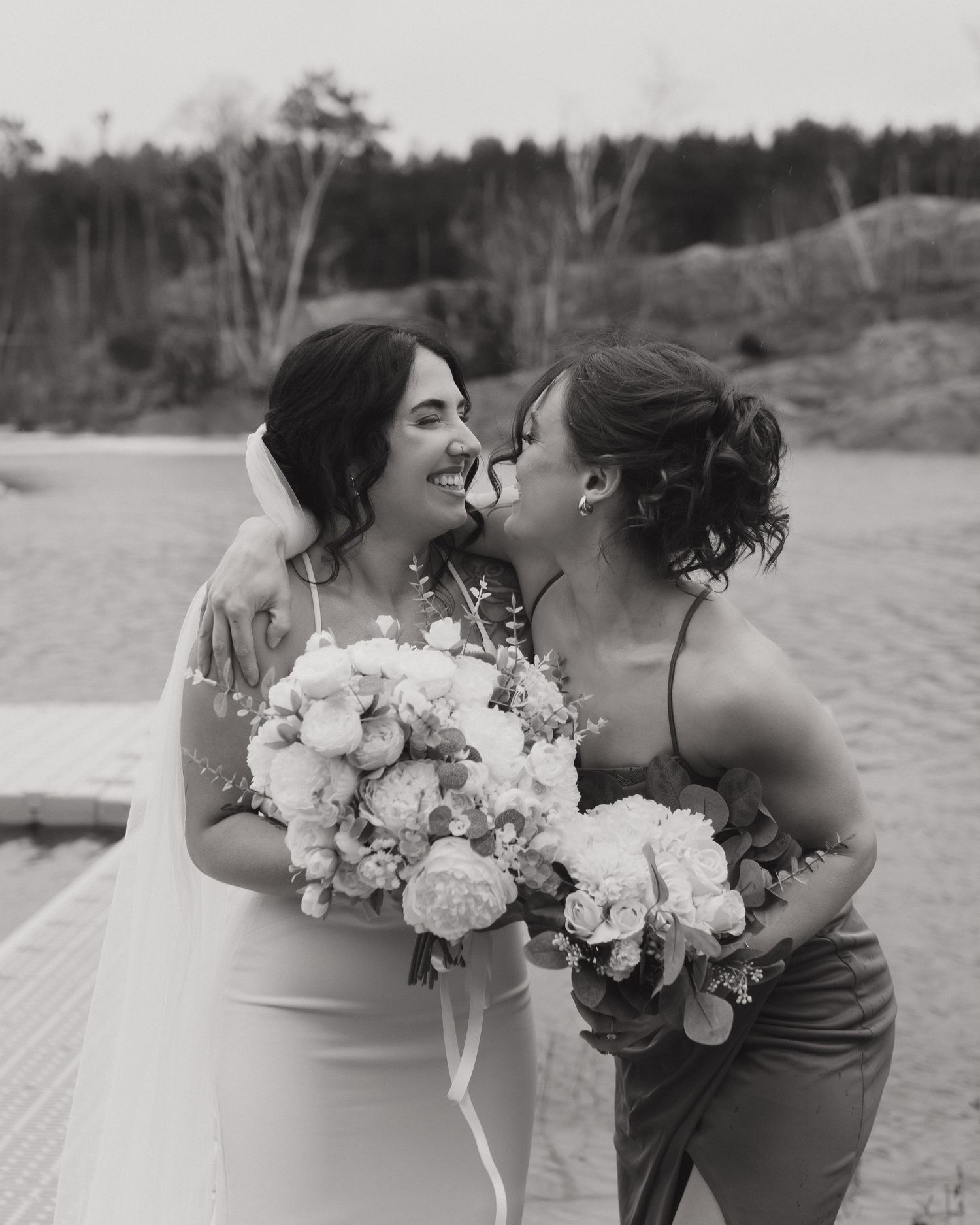 Black and white photo of a bride and bridesmaid laughing together, holding bouquets of peonies at an outdoor wedding.
