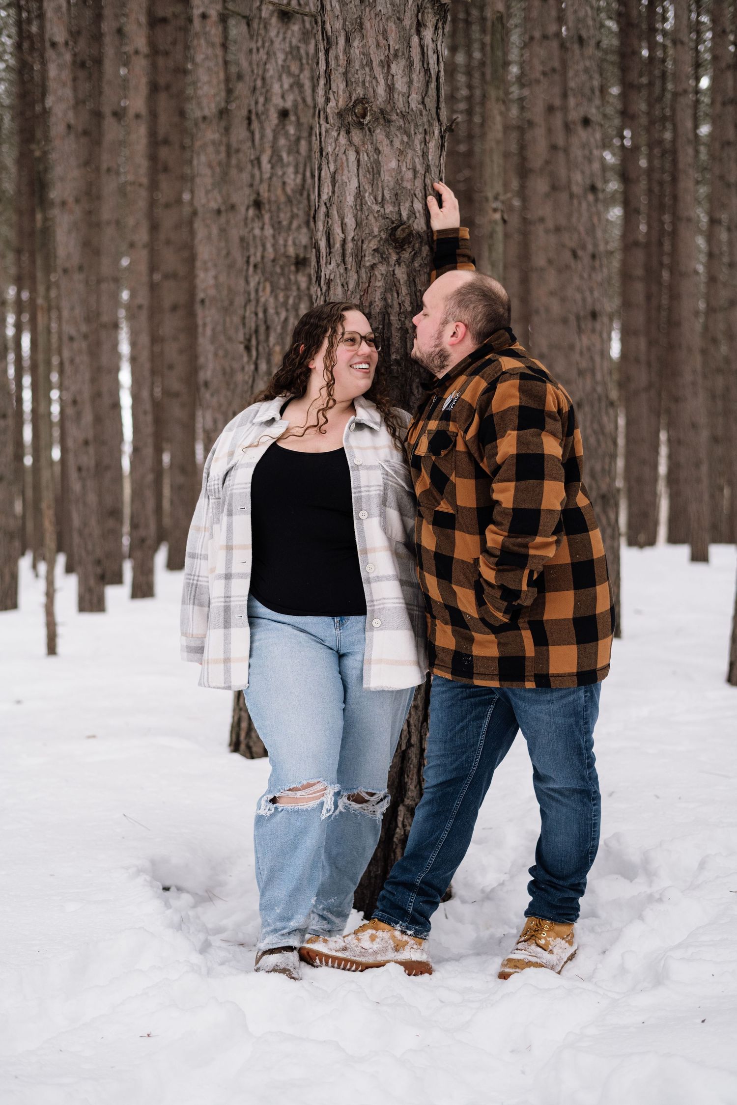Couple in flannel shirts smiling at each other against a pine tree in a snowy winter forest.