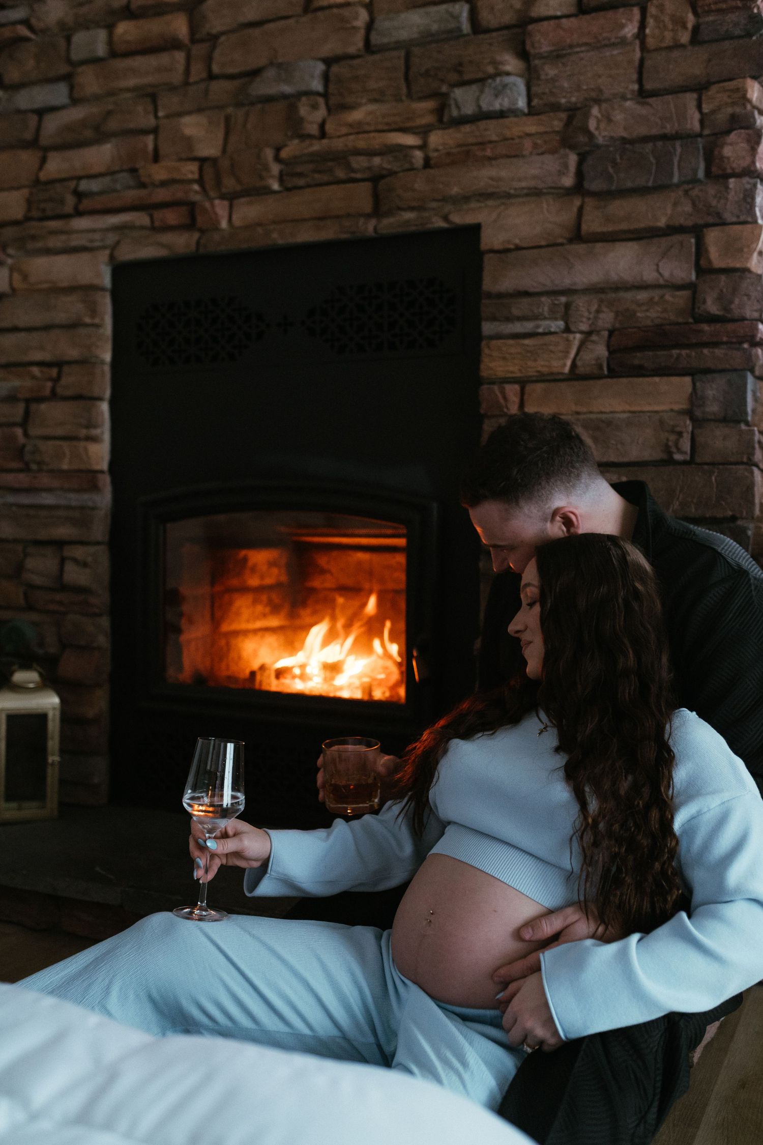 Pregnant woman holding champagne glass with partner by a glowing stone fireplace.