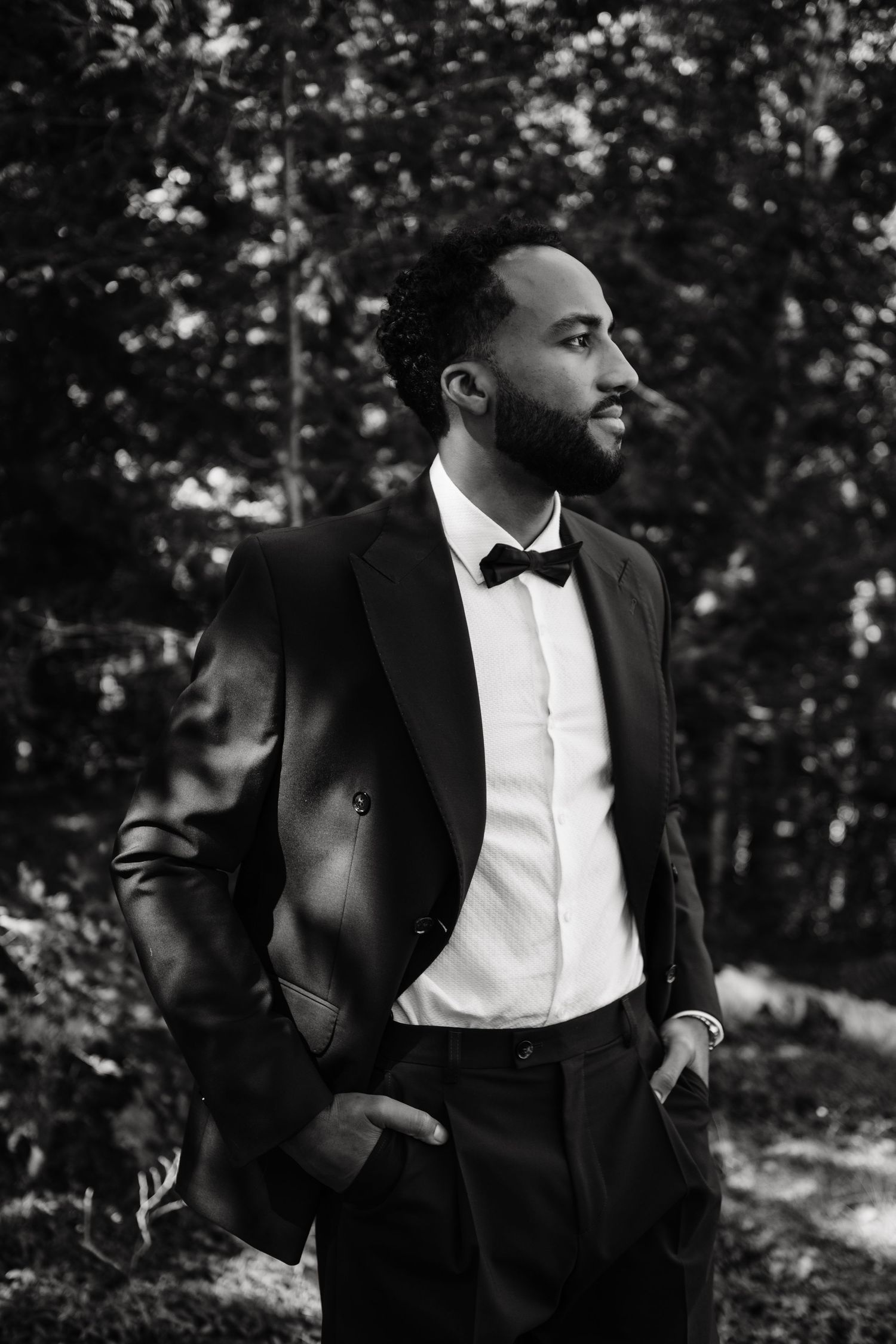 Black and white portrait of a bearded man in a tuxedo with bow tie, hands in pockets, outdoors.
