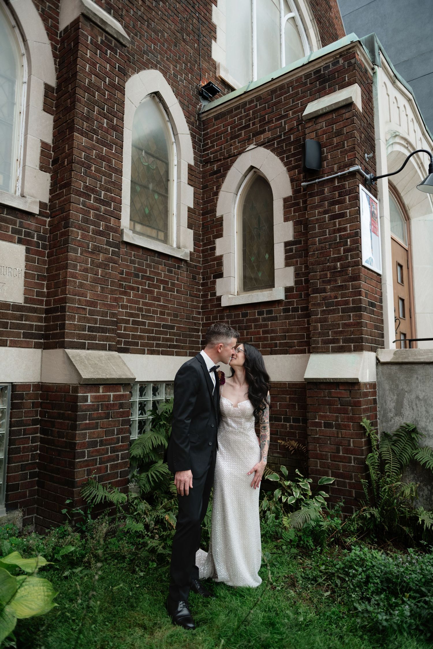 Bride and groom kissing outside a historic brick church, bride in white gown, groom in black suit.