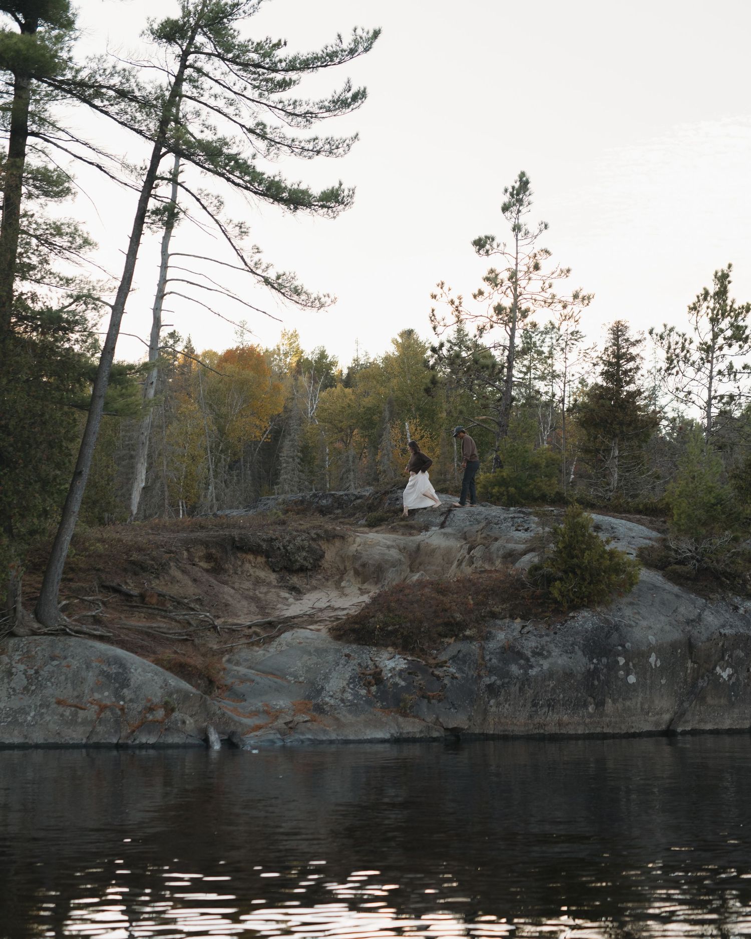 Rocky cliffs and pine trees line the shoreline of a tranquil lake at sunset.