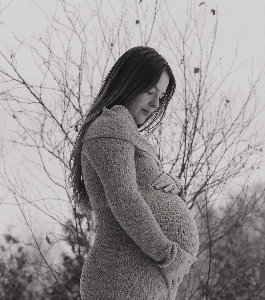 A person in a grey sweater poses against bare winter branches.