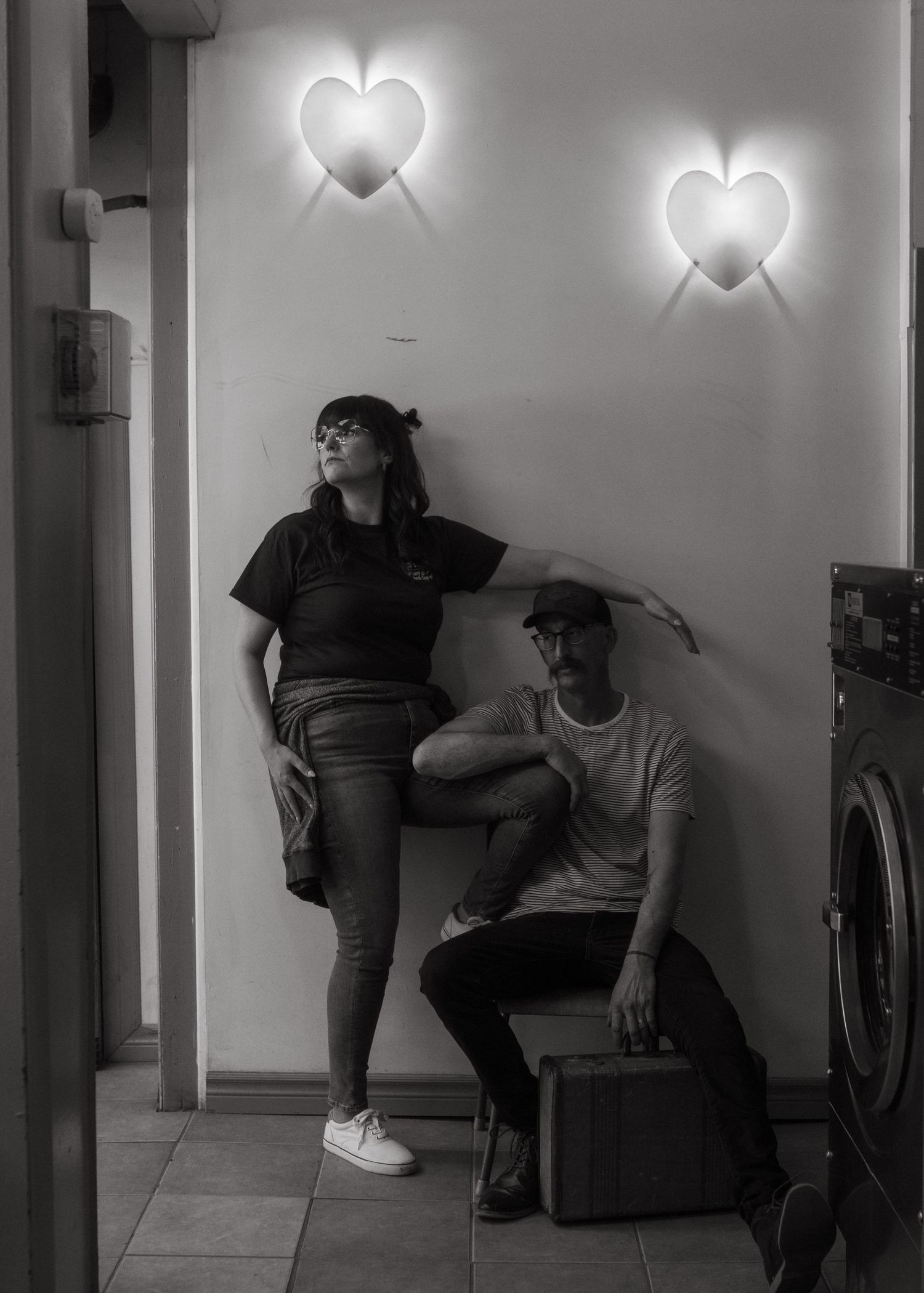 Two people pose against wall with glowing heart-shaped lights in black and white photography.