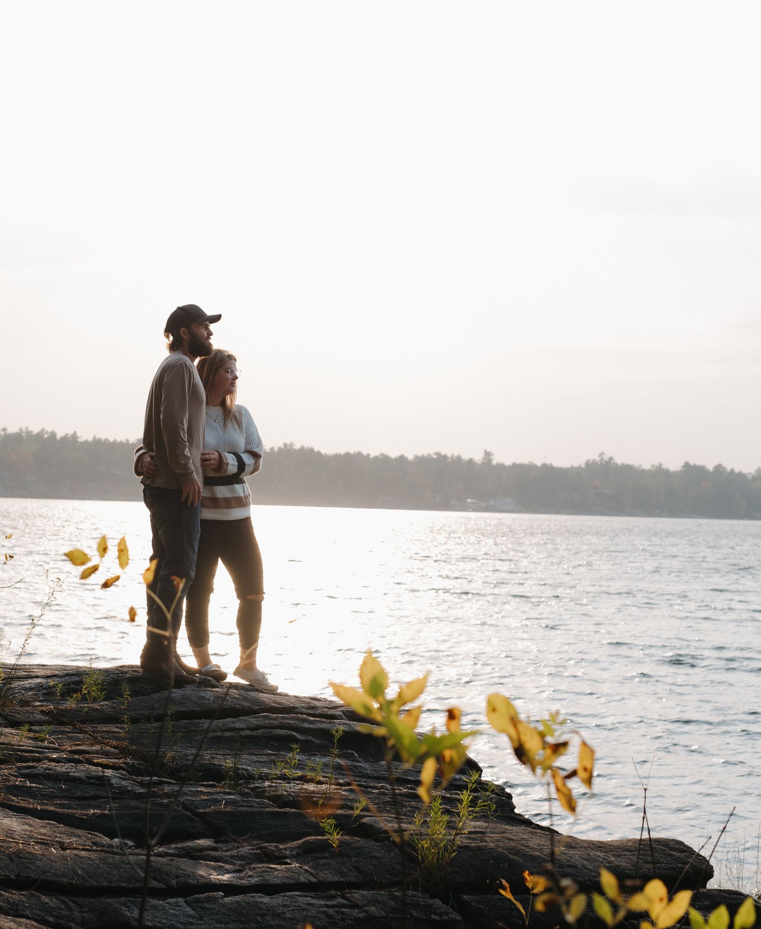 A couple stands silhouetted on rocky shoreline at sunset with yellow flowers in foreground and water views.